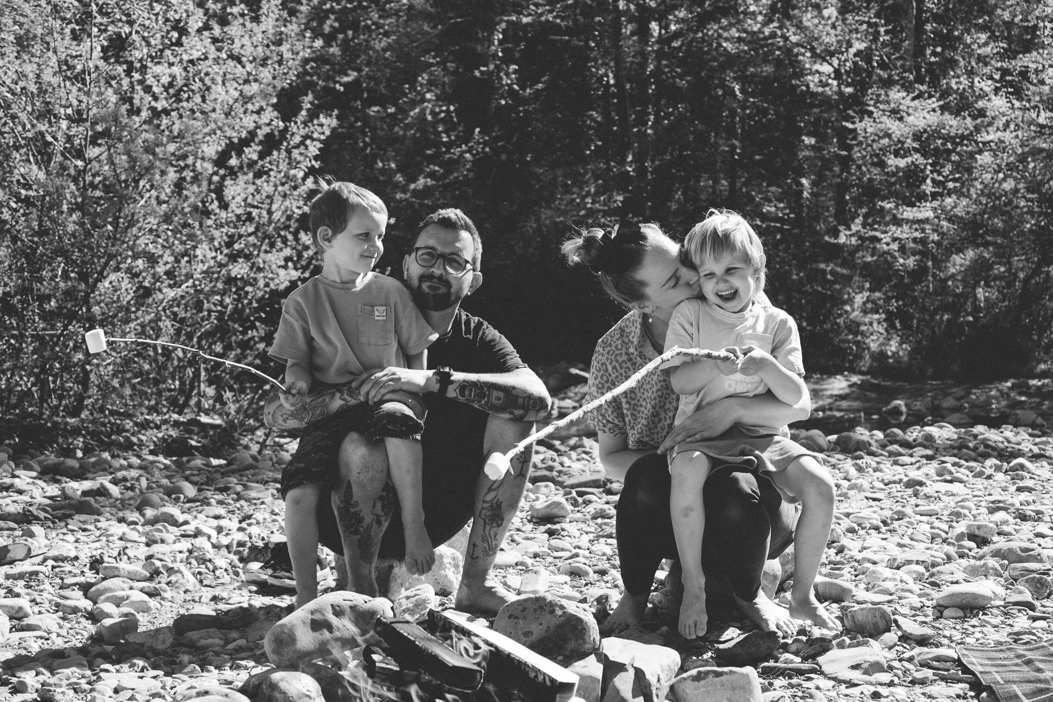 family photoshoot - A black and white photo of a family of four outdoors on a rocky riverbank, with trees in the background. The mother is kissing a young boy on the cheek, while the father and older boy sit nearby, all appearing happy and relaxed.