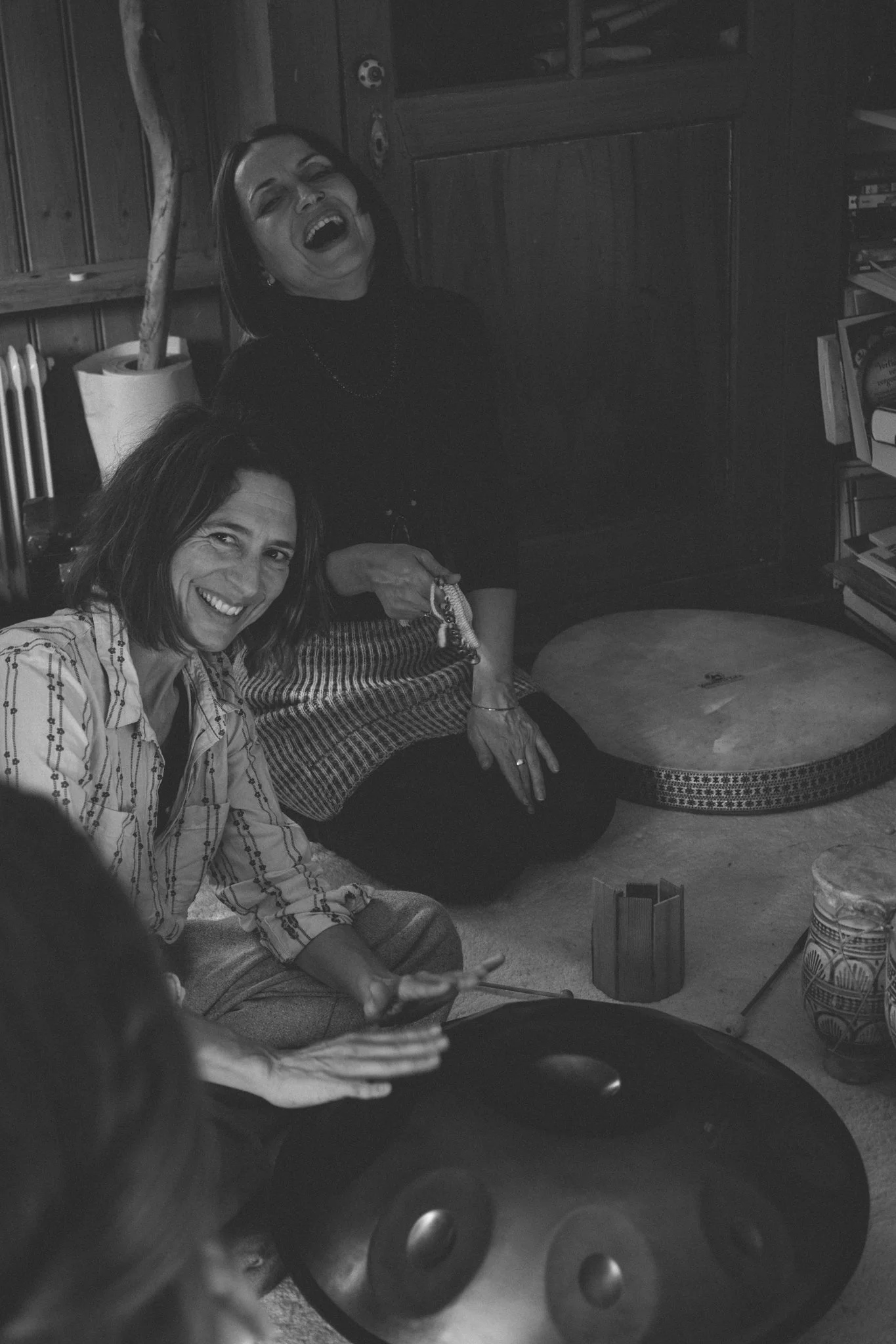 friends photoshoot - Two women sitting on the floor, smiling and laughing. One woman is holding a smartphone. the room has wooden walls, shelves filled with books.