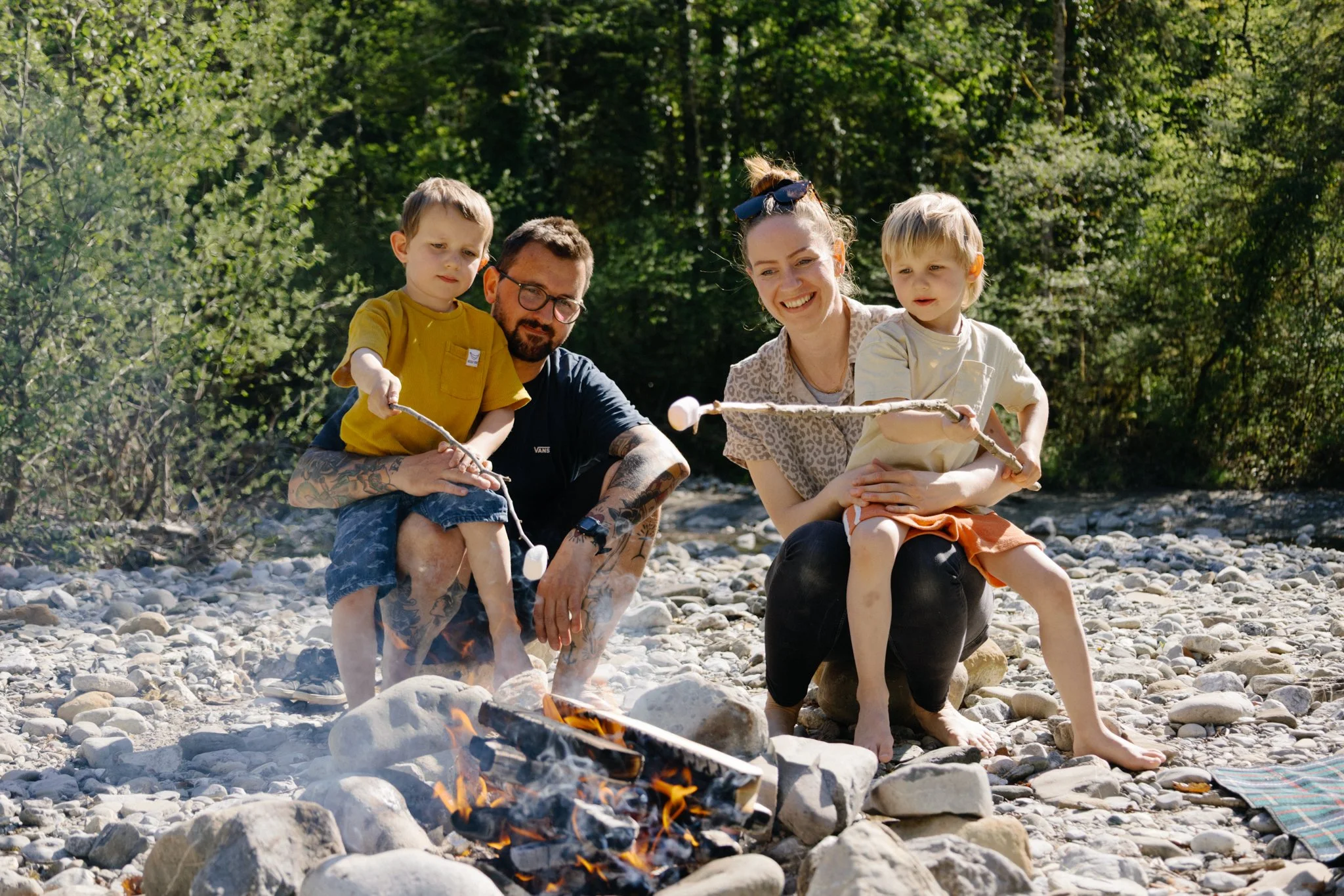 family photoshoot - A family of four sitting on a rocky riverbank beside a campfire, with trees in the background. The adults and children are smiling and holding sticks over the fire.