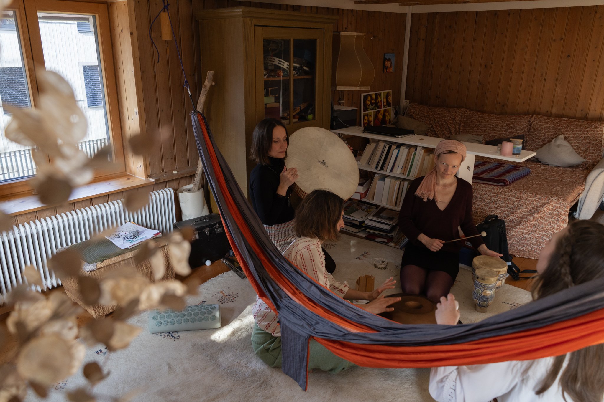 friends photoshoot - A group of women gathered in a cozy wooden room, sitting on the floor and engaging in a musical activity with percussion instruments. The room has wooden walls, a bookshelf filled with books, in a casual, home-like setting.