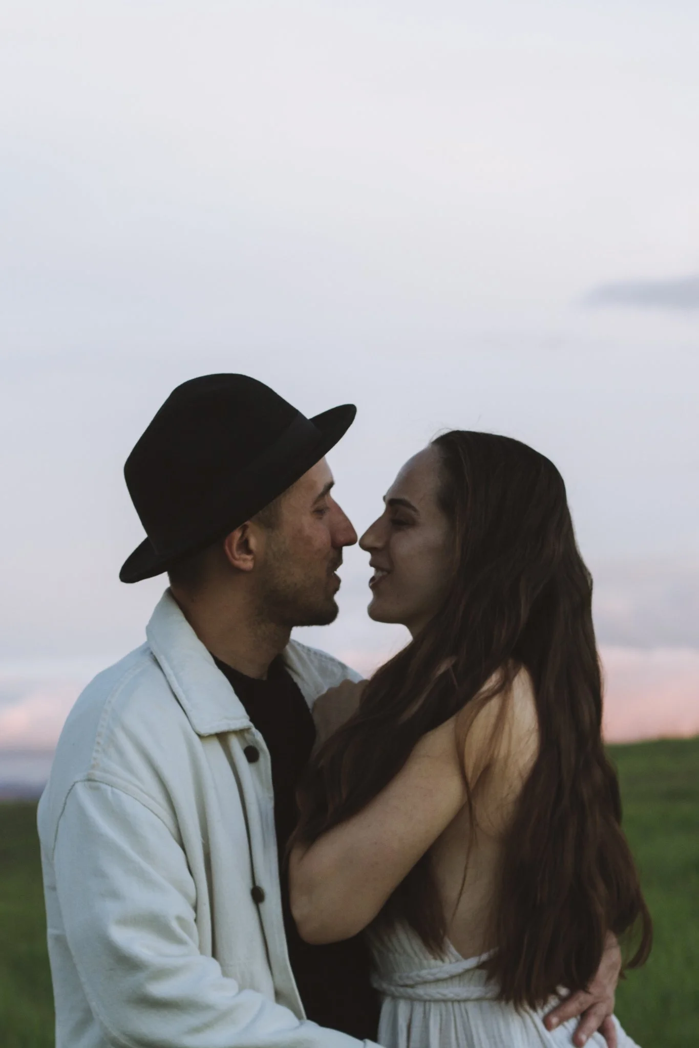 couples photoshoot - A couple stands close together outdoors, gazing into each other's eyes, with a grassy field and cloudy sky in the background.