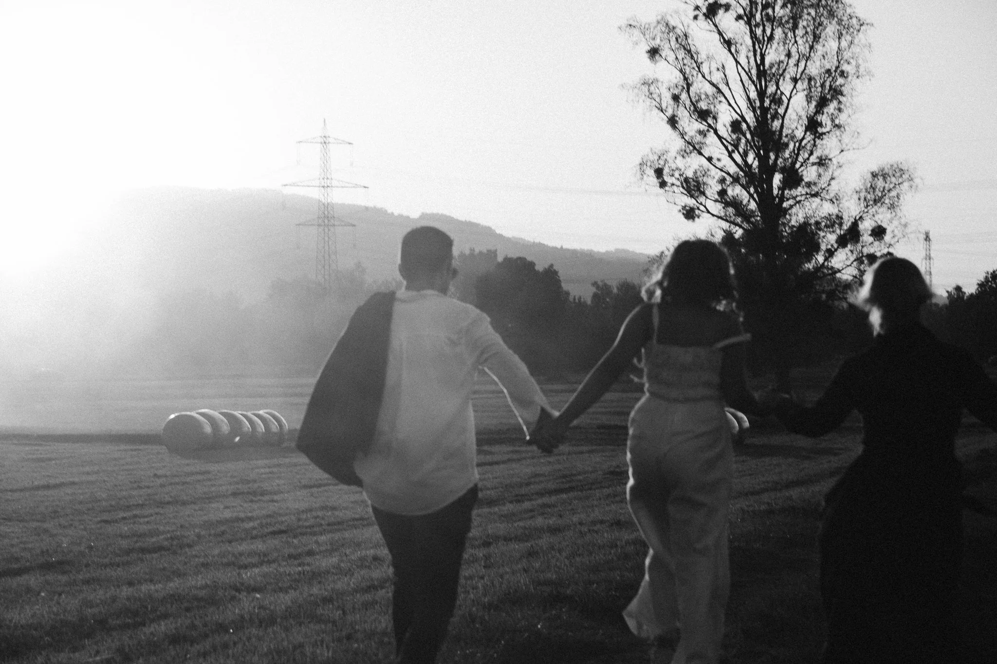family photoshoot - Black and white photo of family offour people holding hands and running outdoors at sunset or sunrise with trees, hills, and power lines in the background.