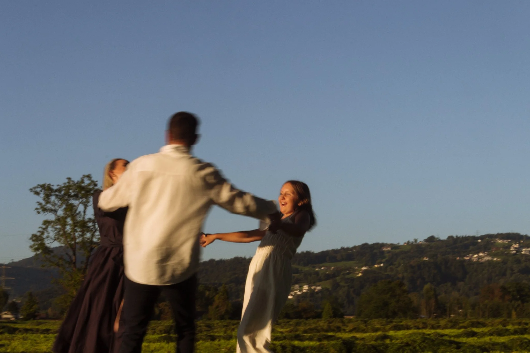 family photoshoot - family dancing outdoors during sunset, smiling, with trees and hills in the background.