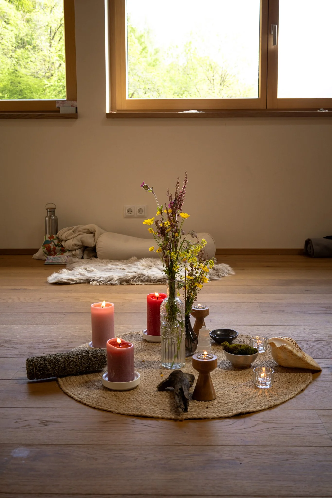 retreat photography - A cozy interior scene with a wooden floor, a circular jute rug with candles, a glass vase with flowers, and decorative items including bowls, stones, and candles on the rug.