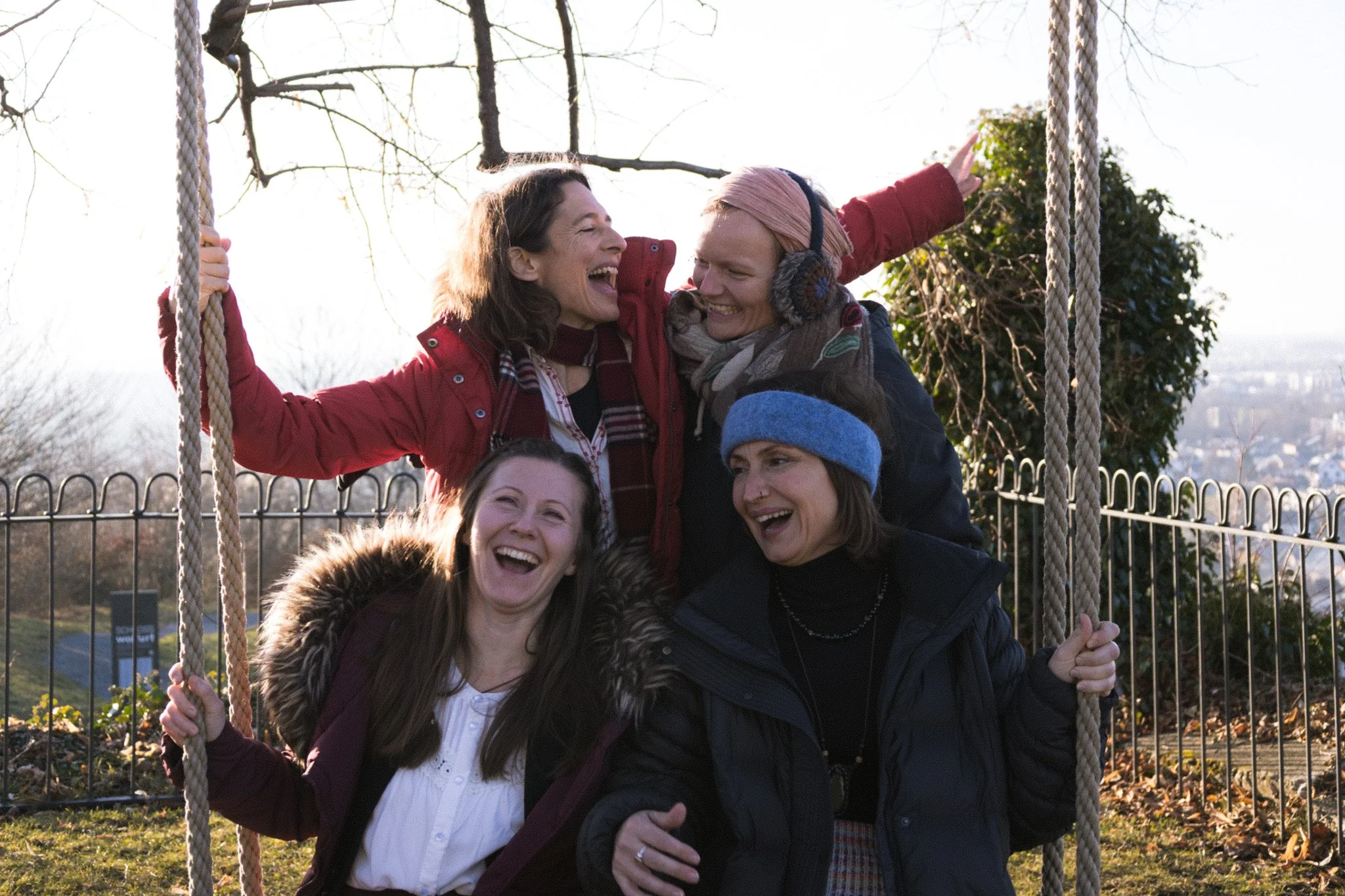 friends photoshoot - A group of four women enjoying themselves on a swing set outdoors, smiling and laughing, in a park with trees and a fence in the background.