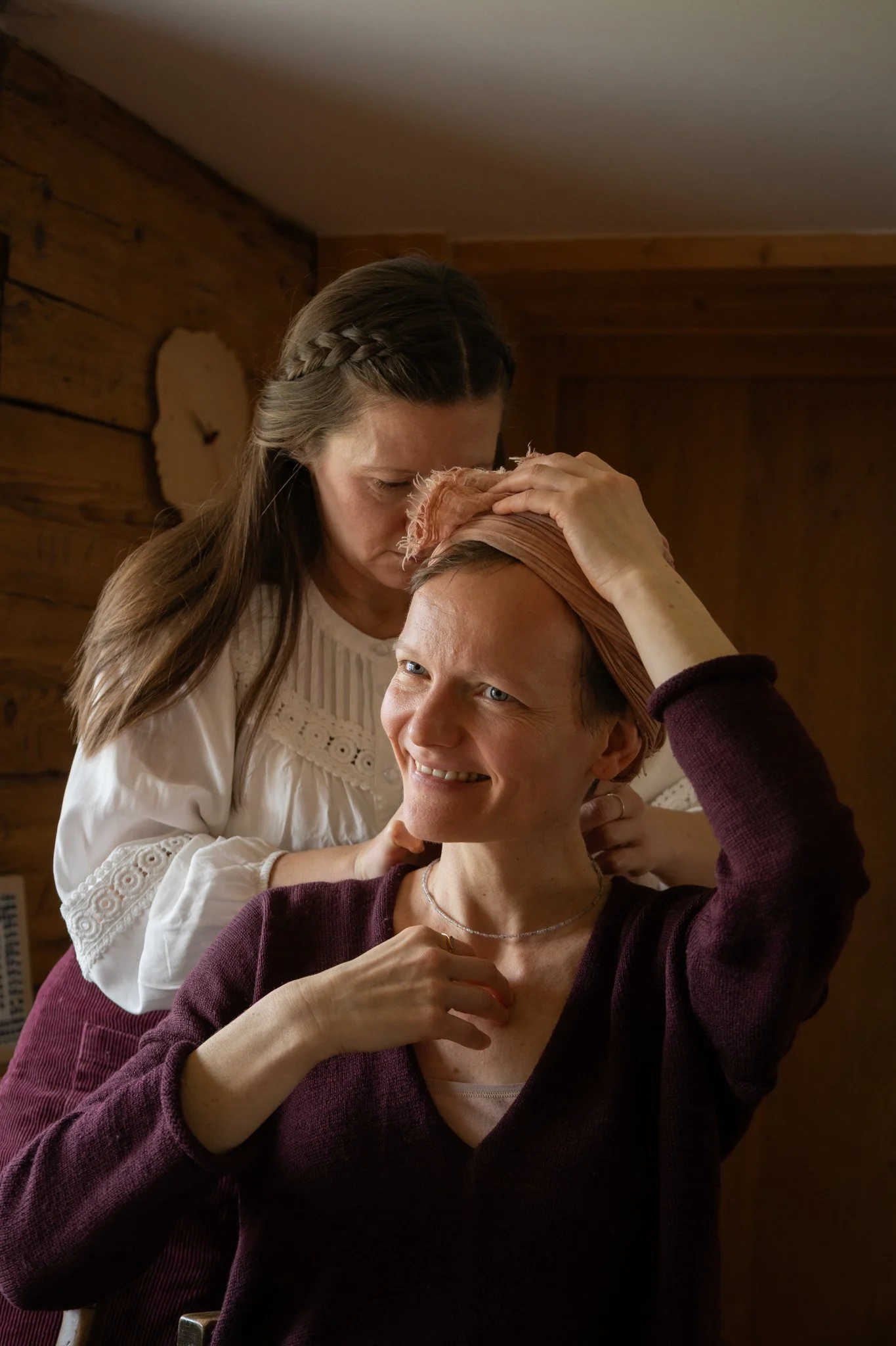 friends photoshoot - A woman with braided hair leaning over another woman with a pink headscarf, who is smiling and wearing a maroon sweater, in a cozy wooden room.