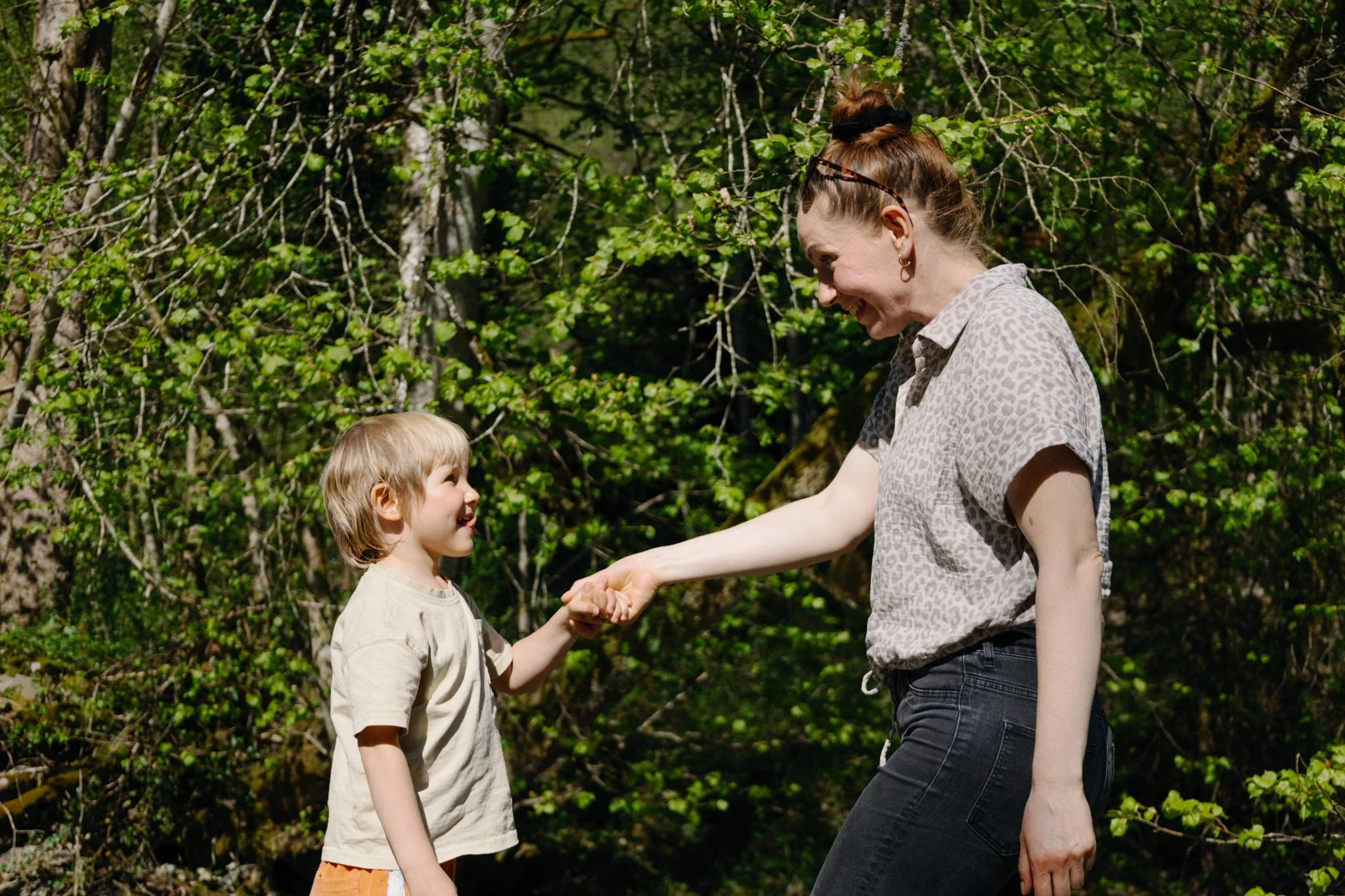 family photoshoot - A woman and a young boy smiling and holding hands outdoors among green trees.