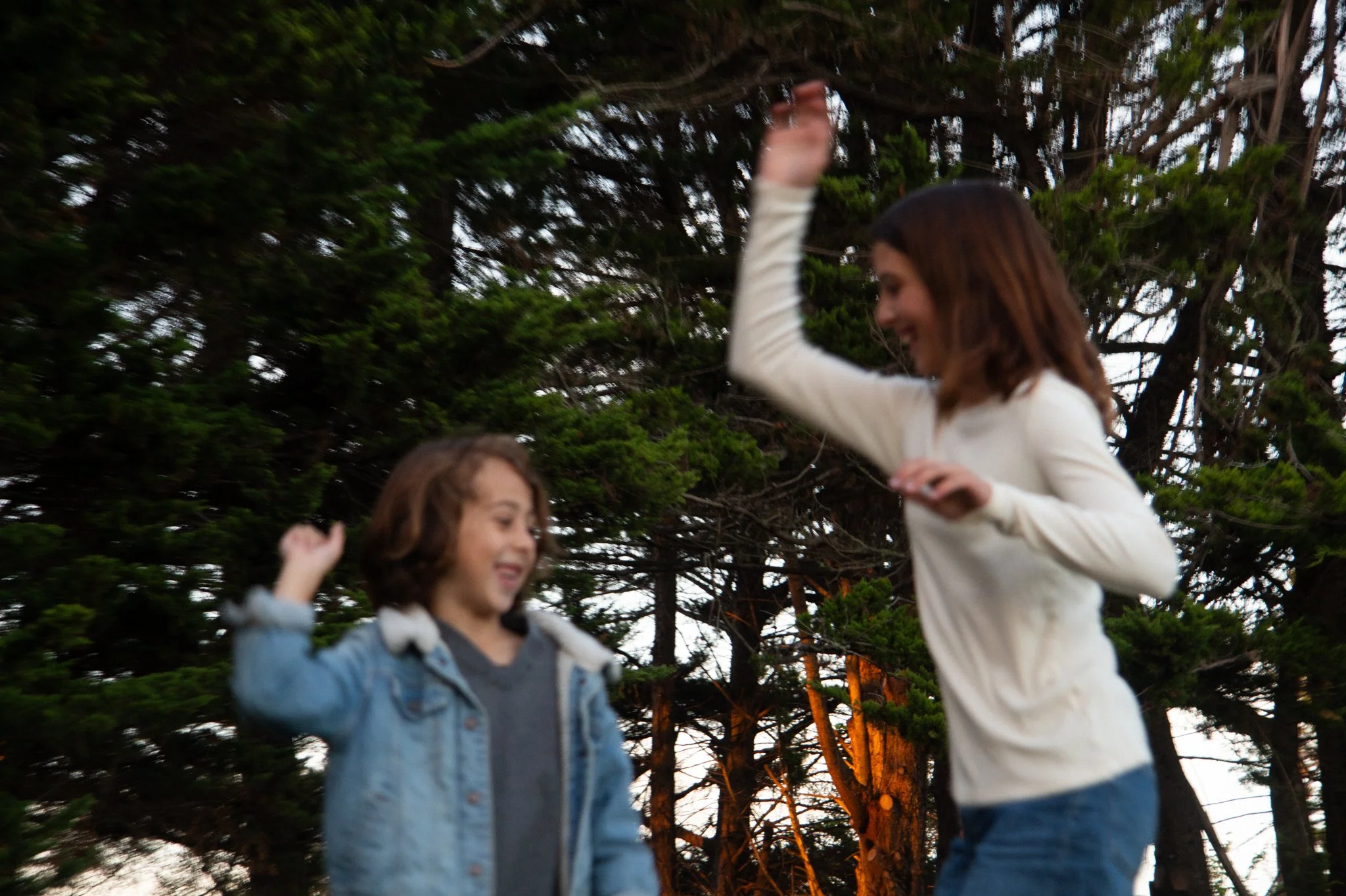 family photo shoot - Two kids outdoors near a large tree, one reaching up and smiling while the other looks down and smiles.