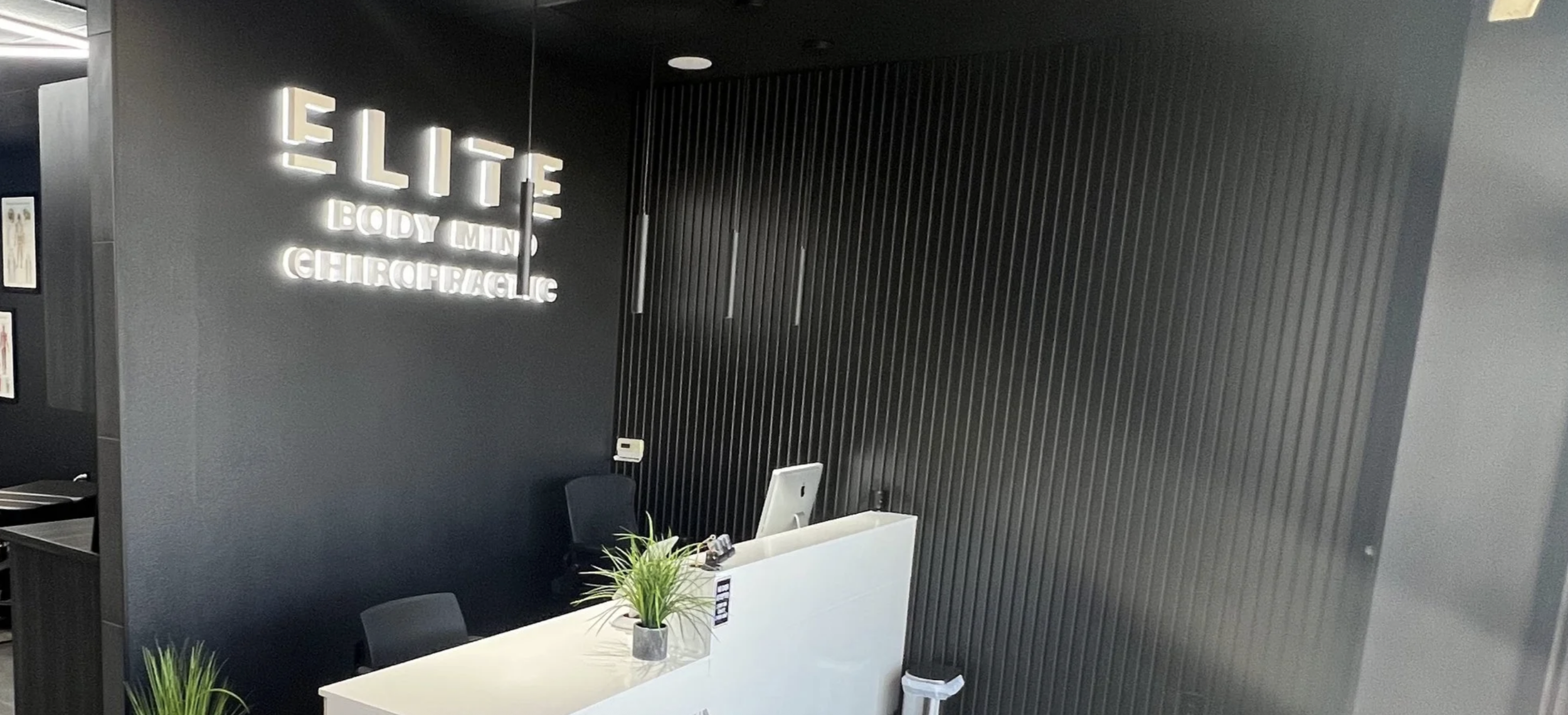 Reception desk at Elite Body Mind Chiropractic Clinic with a potted plant, dark walls, and illuminated sign with the clinic's name.