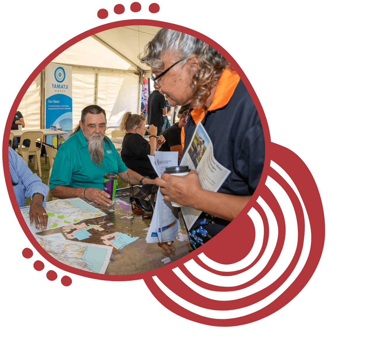 People at an outdoor event inside a tent, sitting at a table with maps, and one person standing holding informational brochures.
