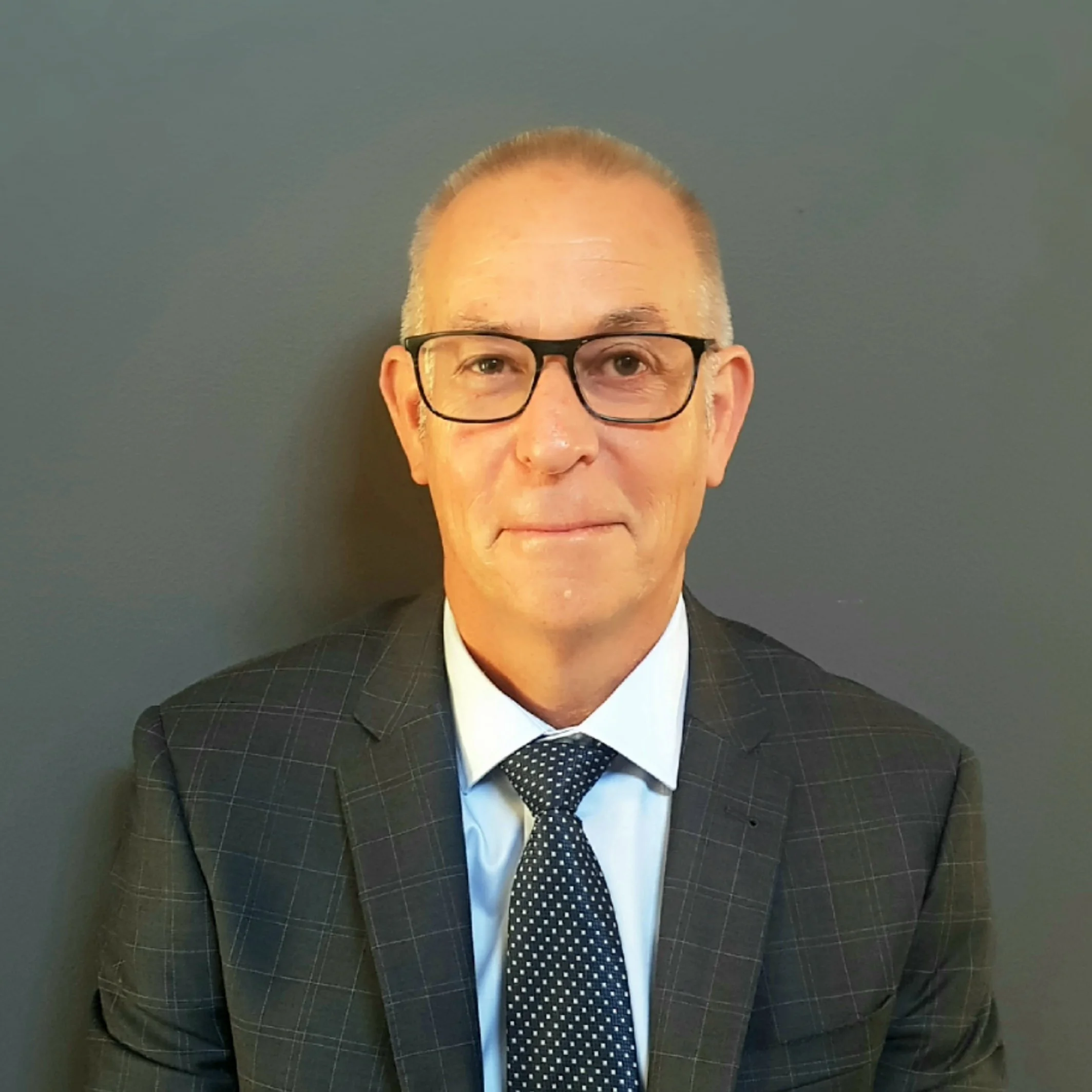 Portrait of a middle-aged man wearing glasses, a dark checkered suit, a white shirt, and a polka dot tie, standing against a plain dark wall.