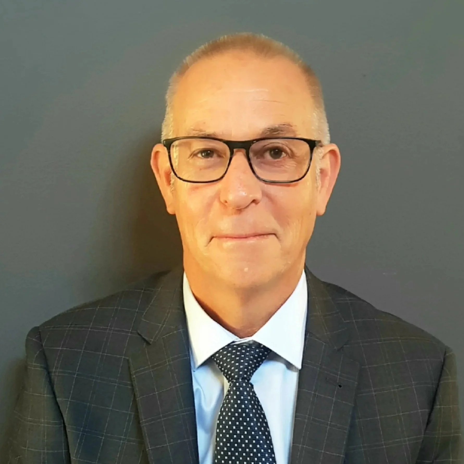 Portrait of a middle-aged man wearing glasses, a dark checkered suit, a white shirt, and a polka dot tie, standing against a plain dark wall.