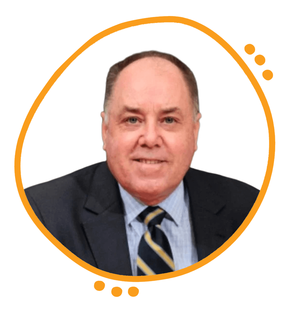 A middle-aged man with light skin, short light brown hair, and blue eyes, wearing a dark suit jacket, light blue shirt, and a striped tie, outdoors with trees in the background.