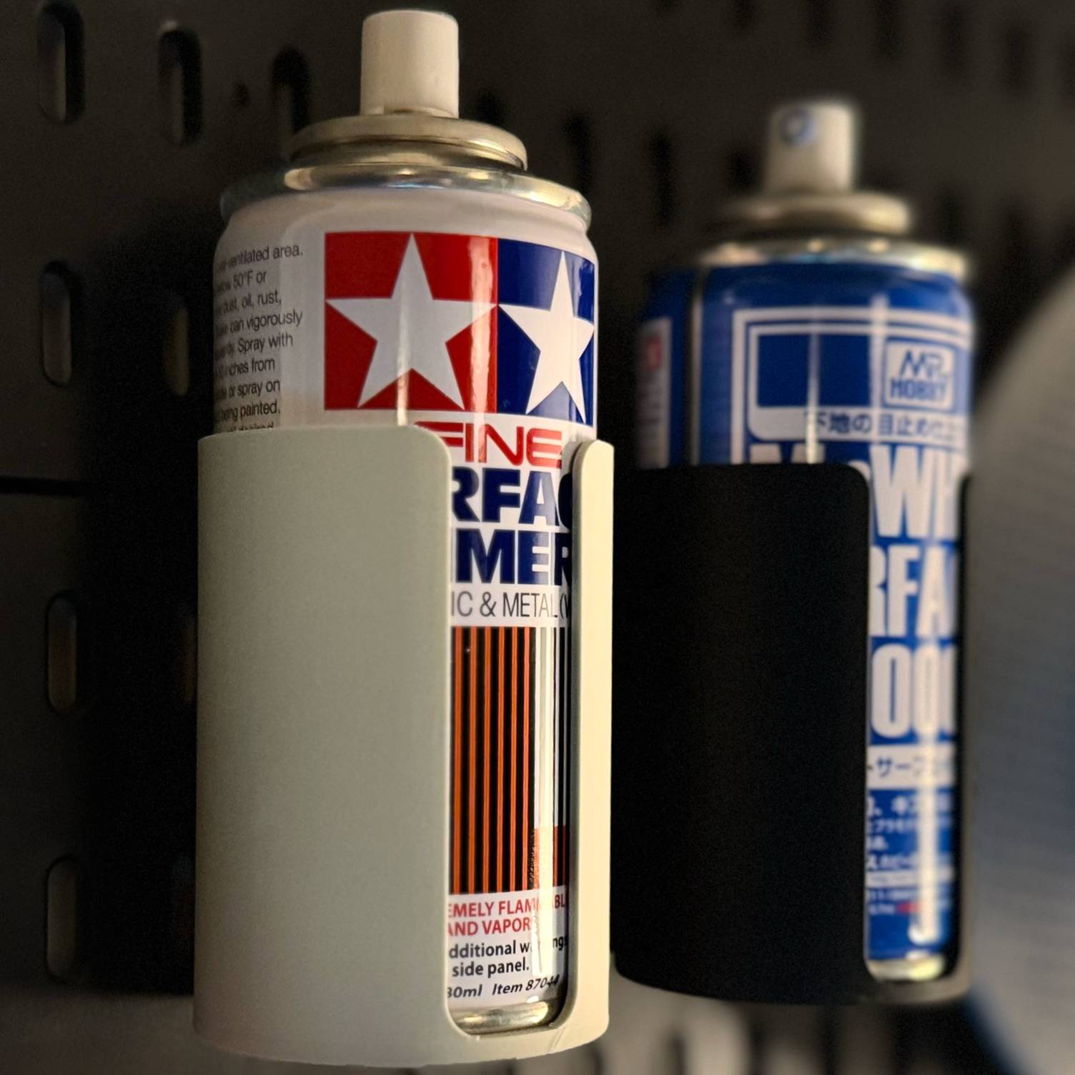 Close-up of two spray cans, one labeled 'Tamiya Fine Surface Primer' with a red, white, and blue star design, mounted on a black pegboard with a gray and black plastic holder, and the other partially visible in the background with a blue and white design.