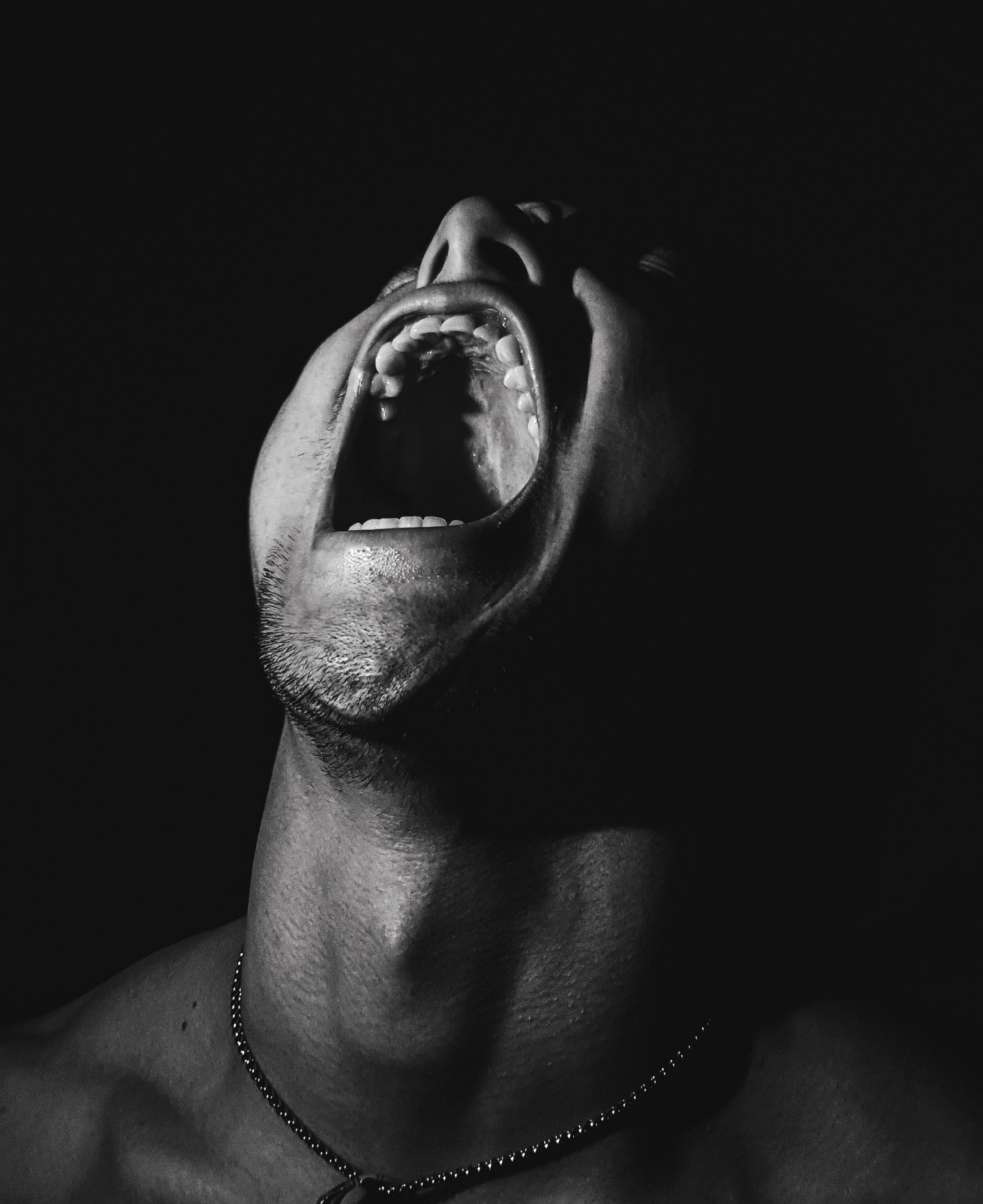 Black and white photo of a man shouting with his mouth wide open against a dark background.