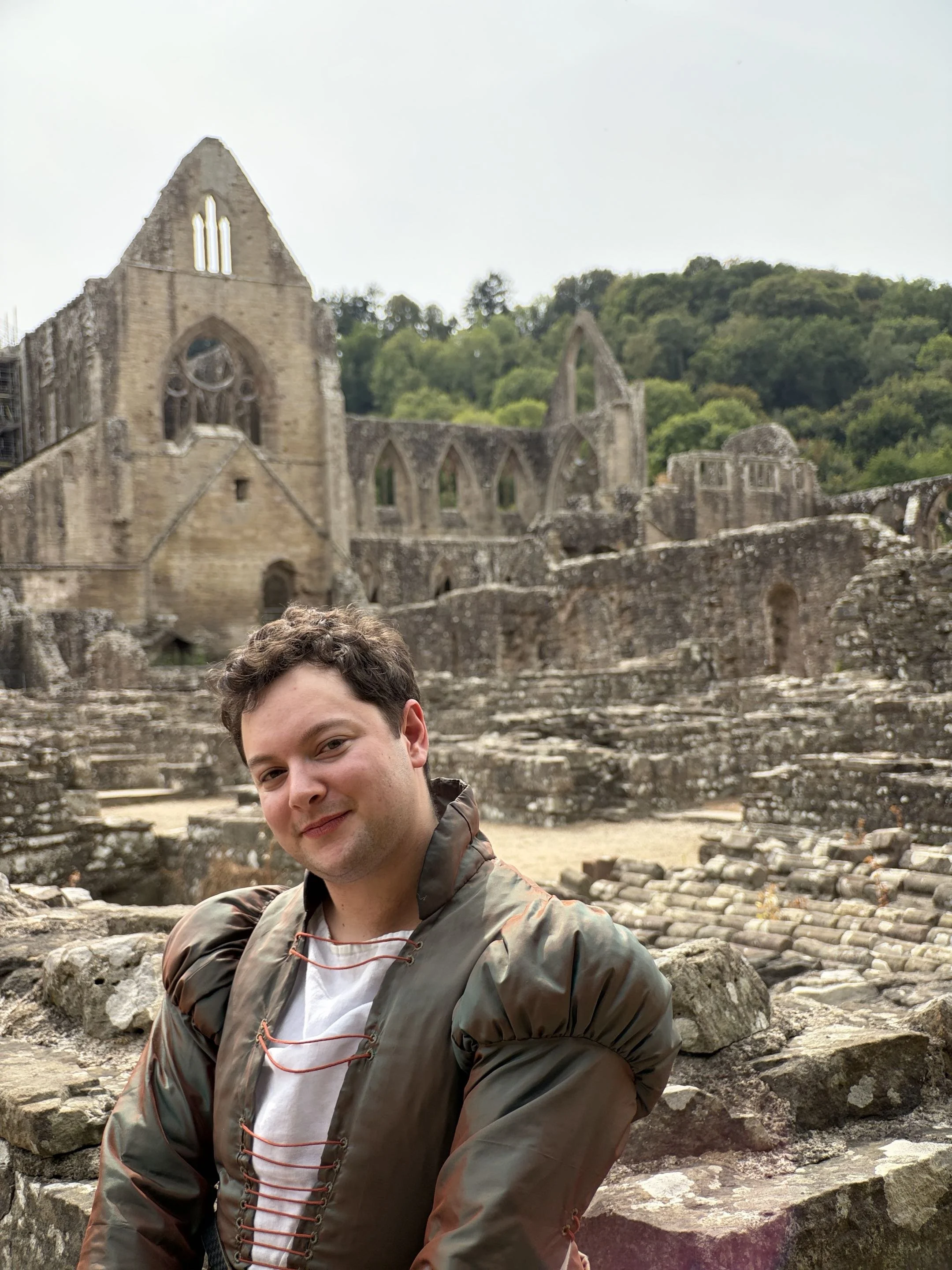 Henric, an androgynous white person with short brown curly hair, smiling at the camera. They are wearing a green and red shot silk doublet, and are sitting on a ruined wall with an abbey in the background.