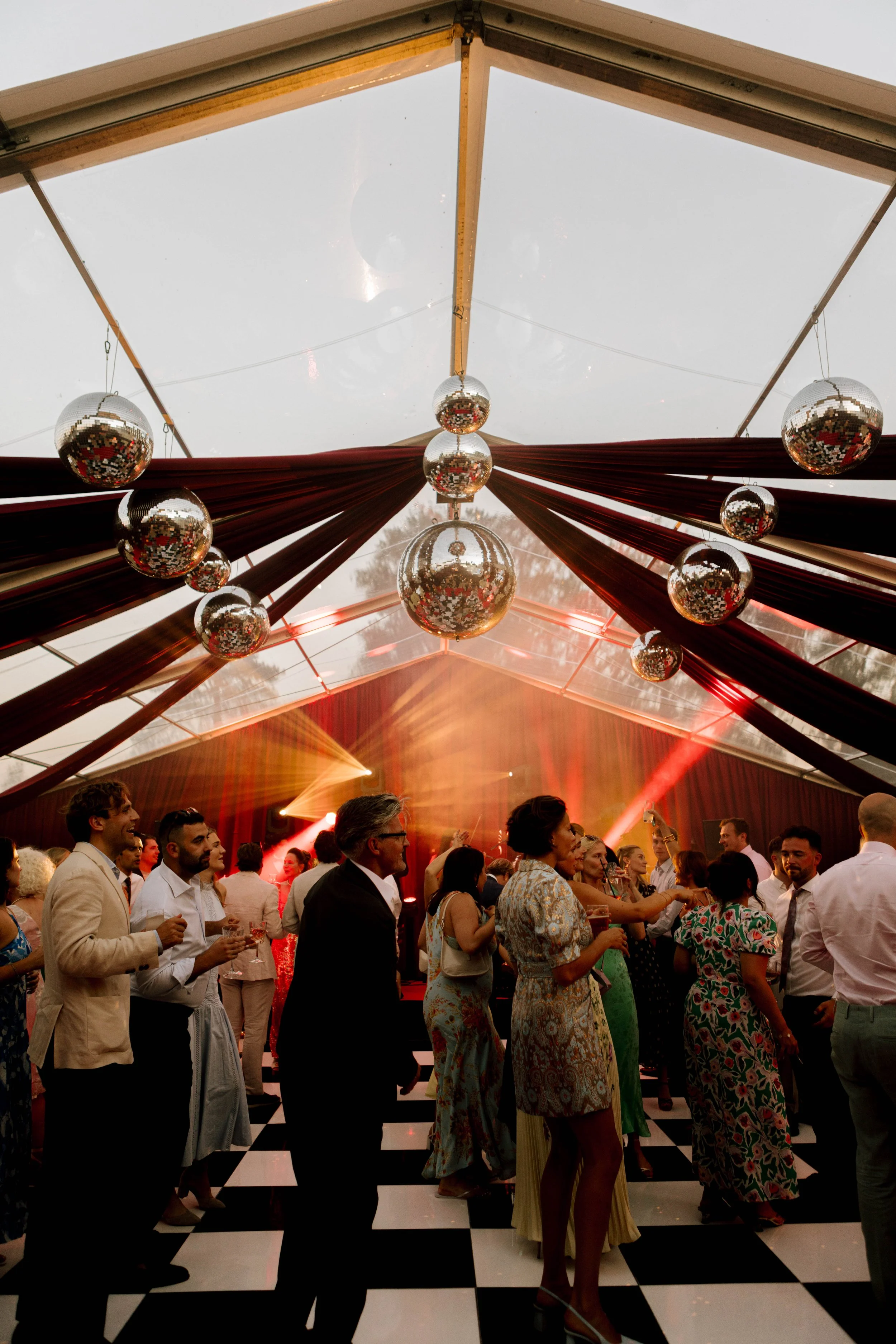 People dancing at a party inside a decorated venue with a black and white checkered floor, draped red fabric, disco balls hanging from the ceiling, and lighting effects.