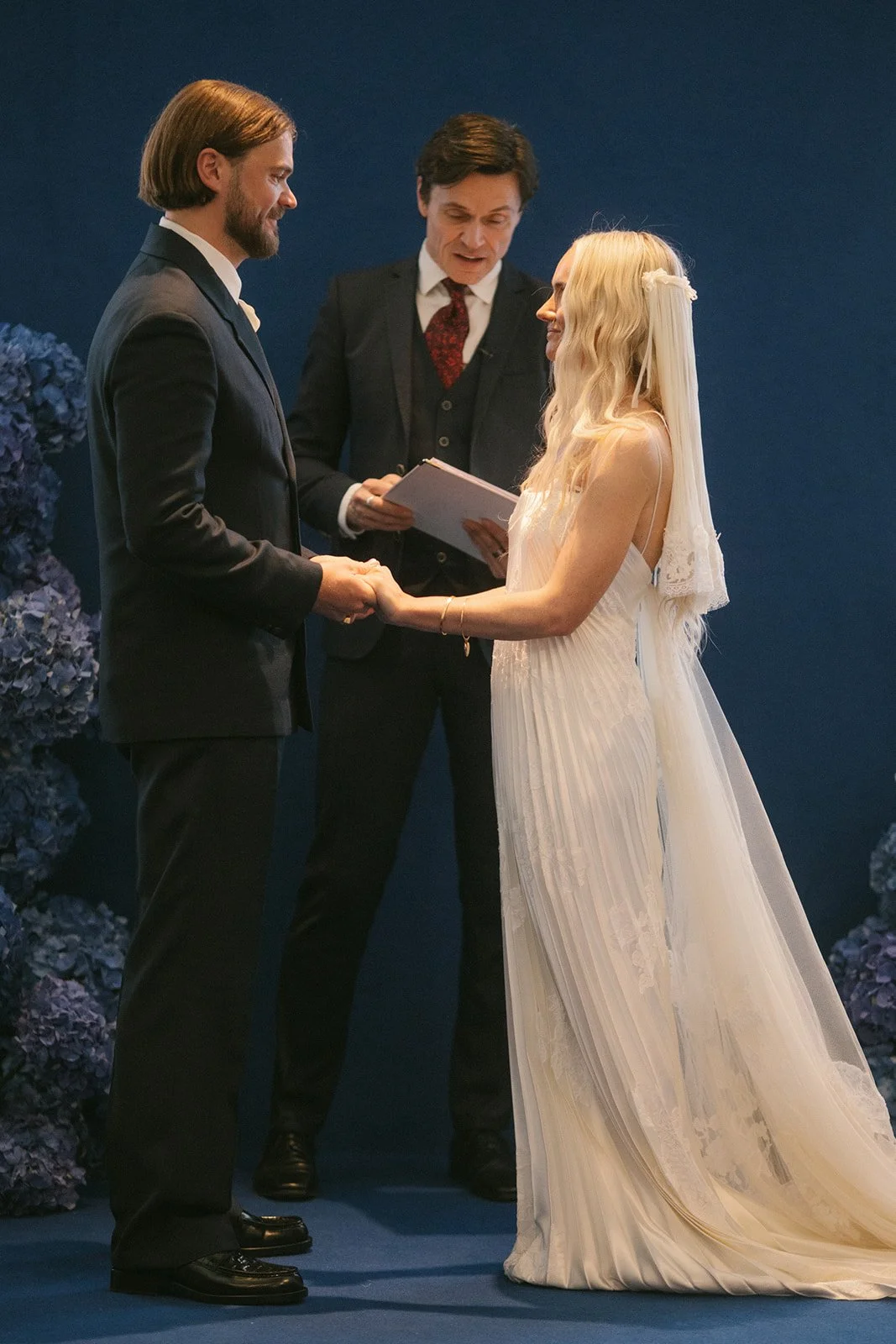A wedding ceremony with a couple holding hands and facing each other, a officiant standing behind them holding a book, and a dark blue background with purple hyacinth flowers on the side.