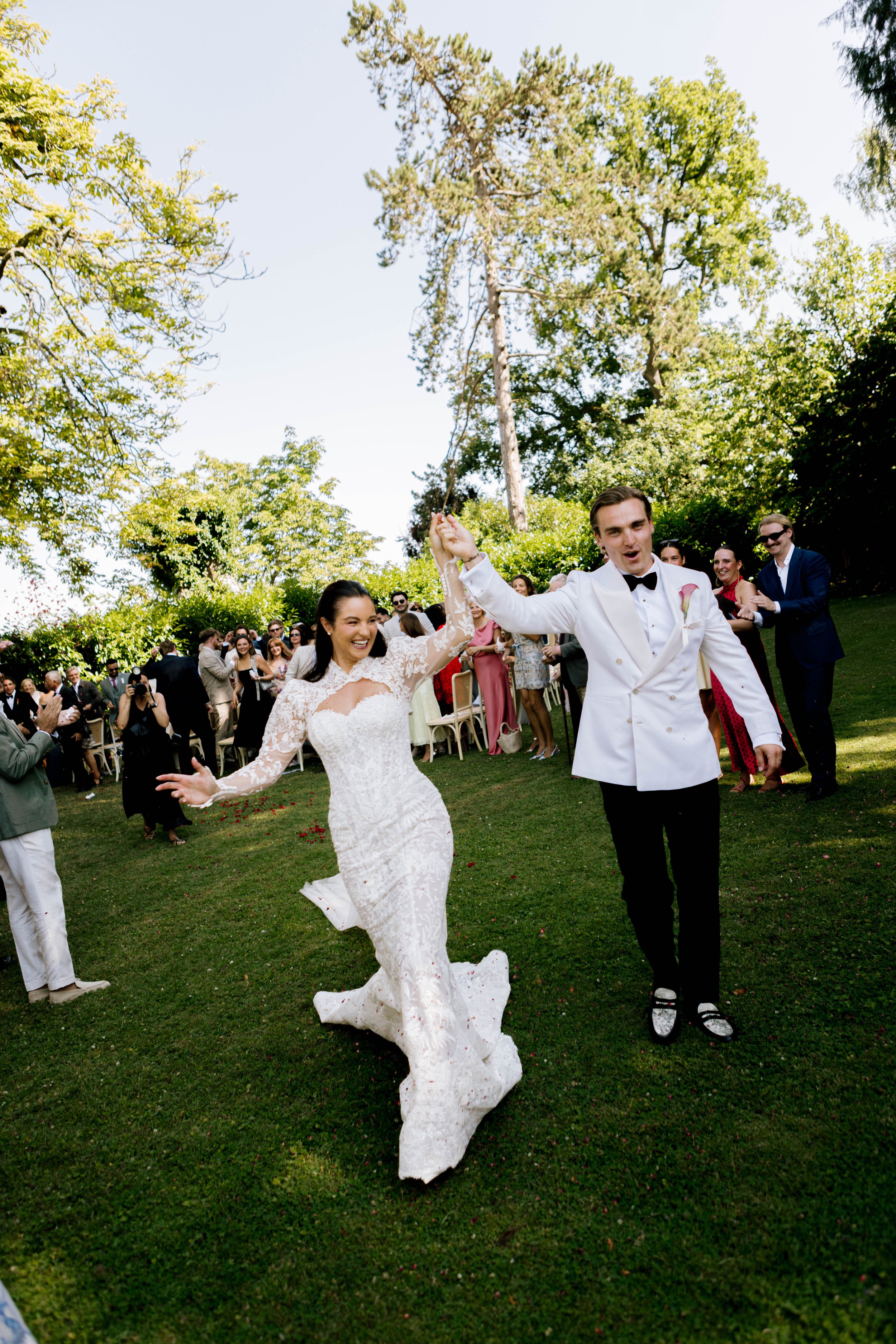 A bride and groom are holding hands and dancing during their wedding reception outdoors. They are surrounded by guests, trees, and a clear blue sky.