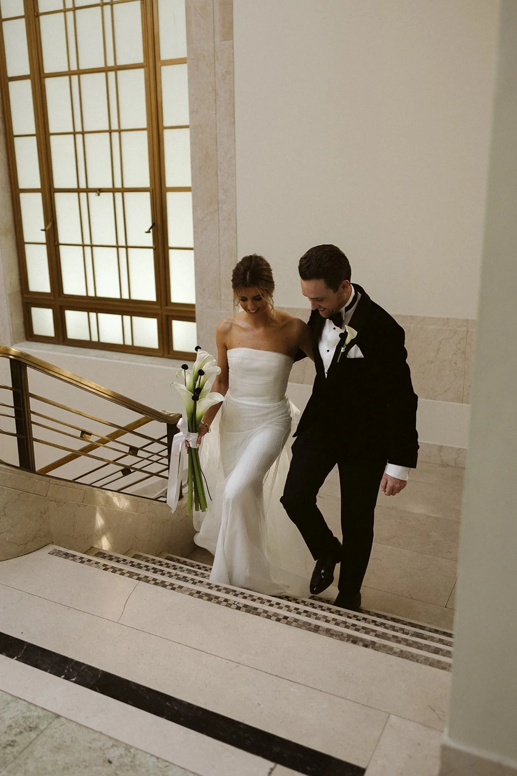 A bride and groom walking up a staircase in an elegant building. The bride is holding a bouquet of calla lilies, and the groom is dressed in a black tuxedo with a white shirt and bow tie.