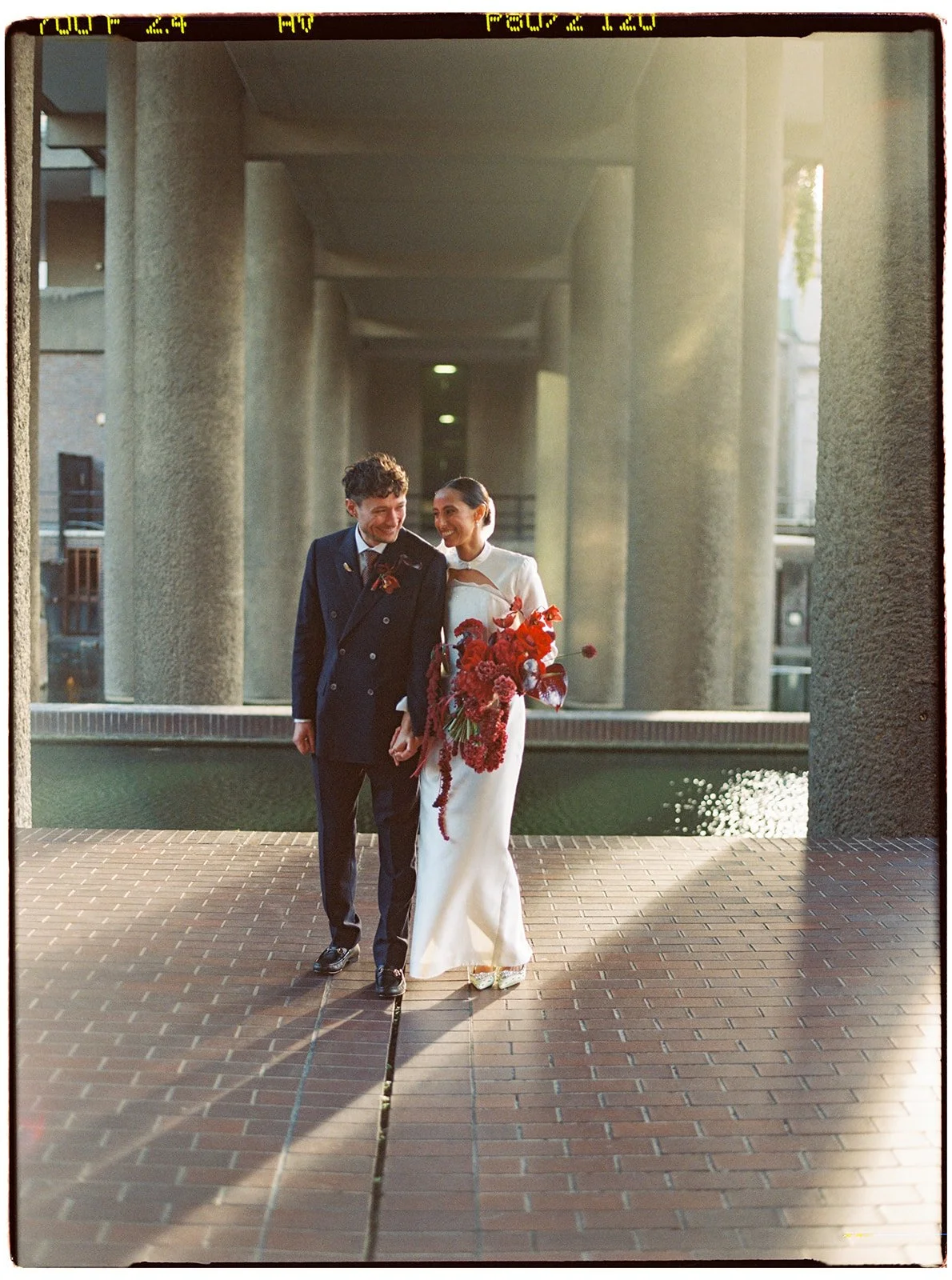 A newly married couple walking hand-in-hand on a brick pathway under a modern concrete bridge. The groom is wearing a dark double-breasted suit and the bride is in a white wedding dress holding a large bouquet of red flowers. They are smiling and loo