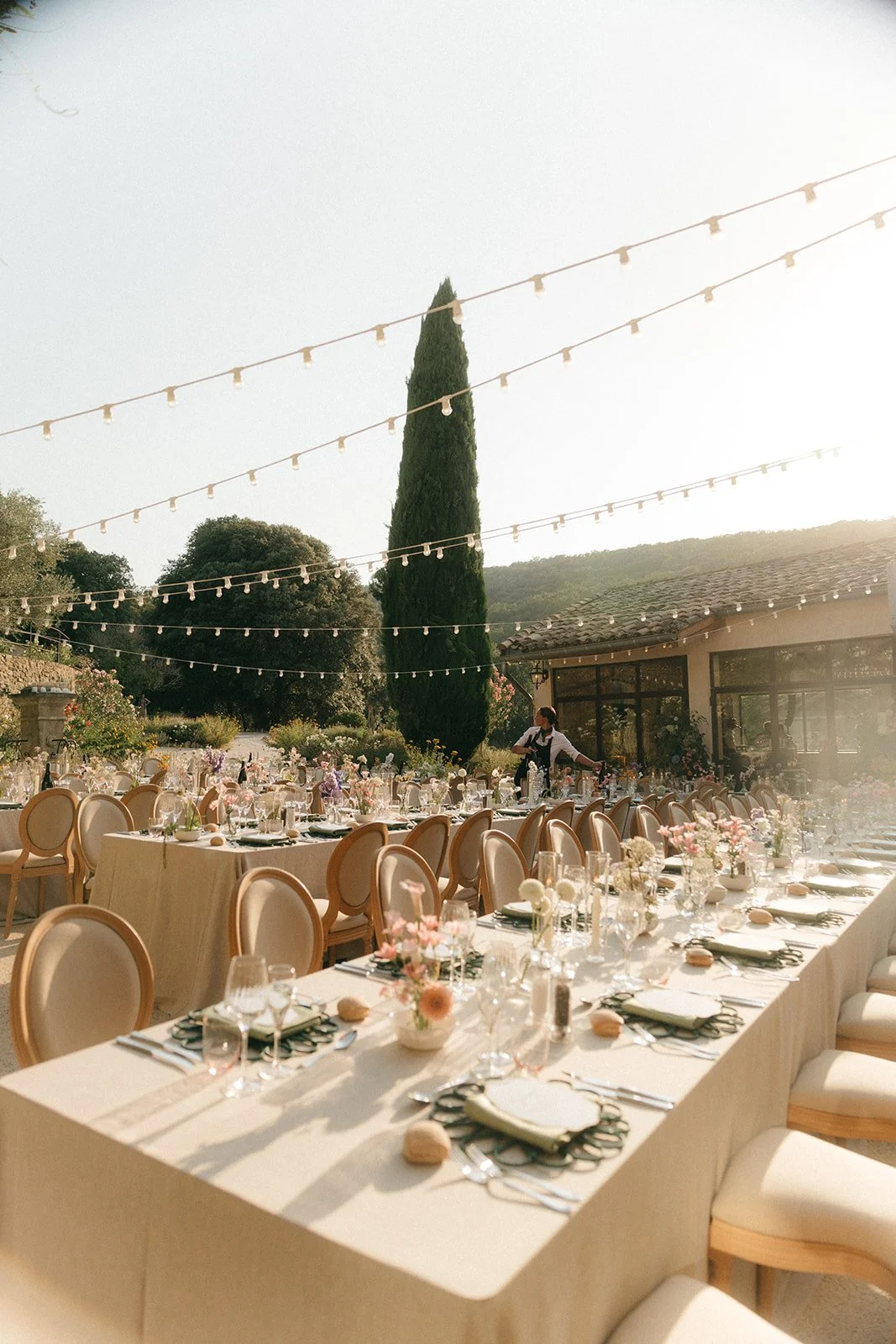 Outdoor banquet setup with long tables decorated with flowers and place settings, string lights hanging above, trees and a building in the background.