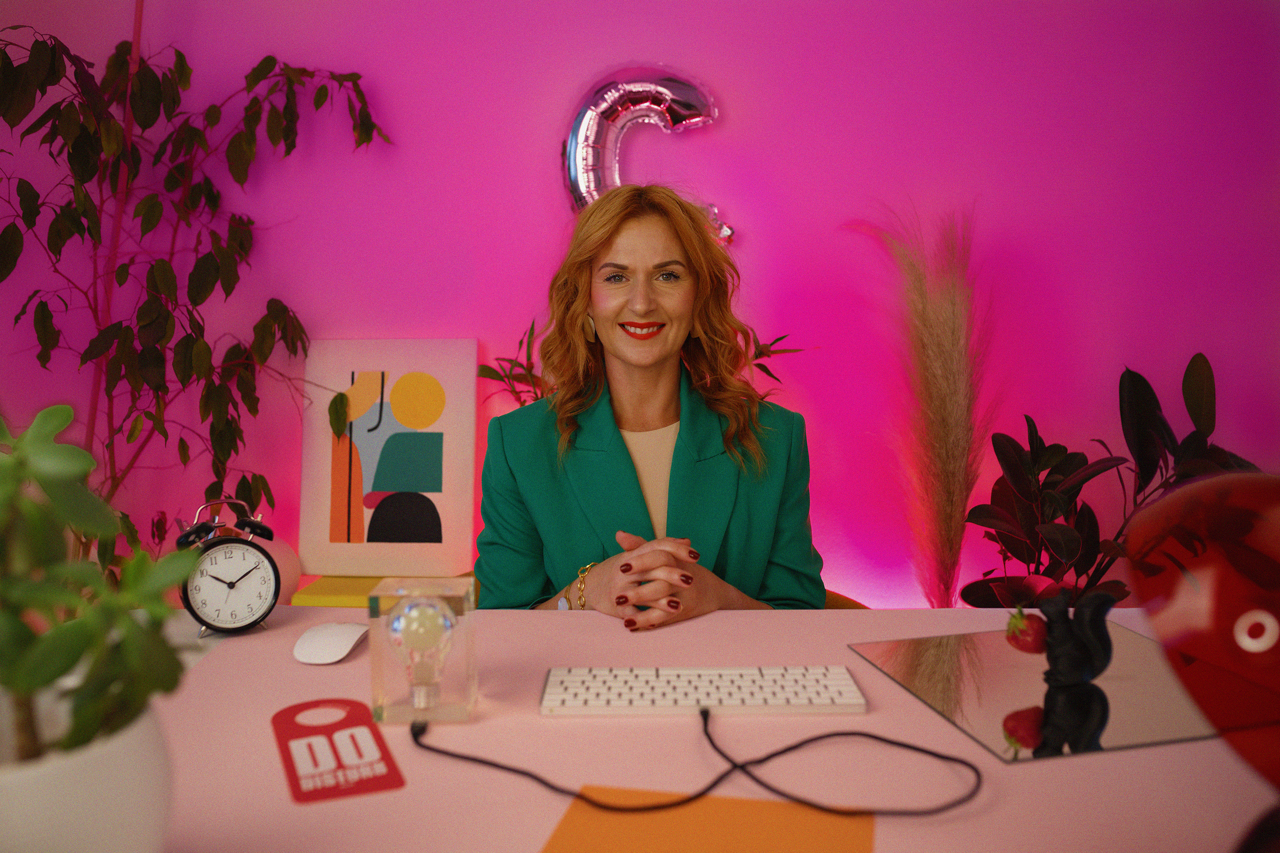 A woman with red hair, wearing a green blazer, sitting at a pink desk with a pink background, smiling at the camera.