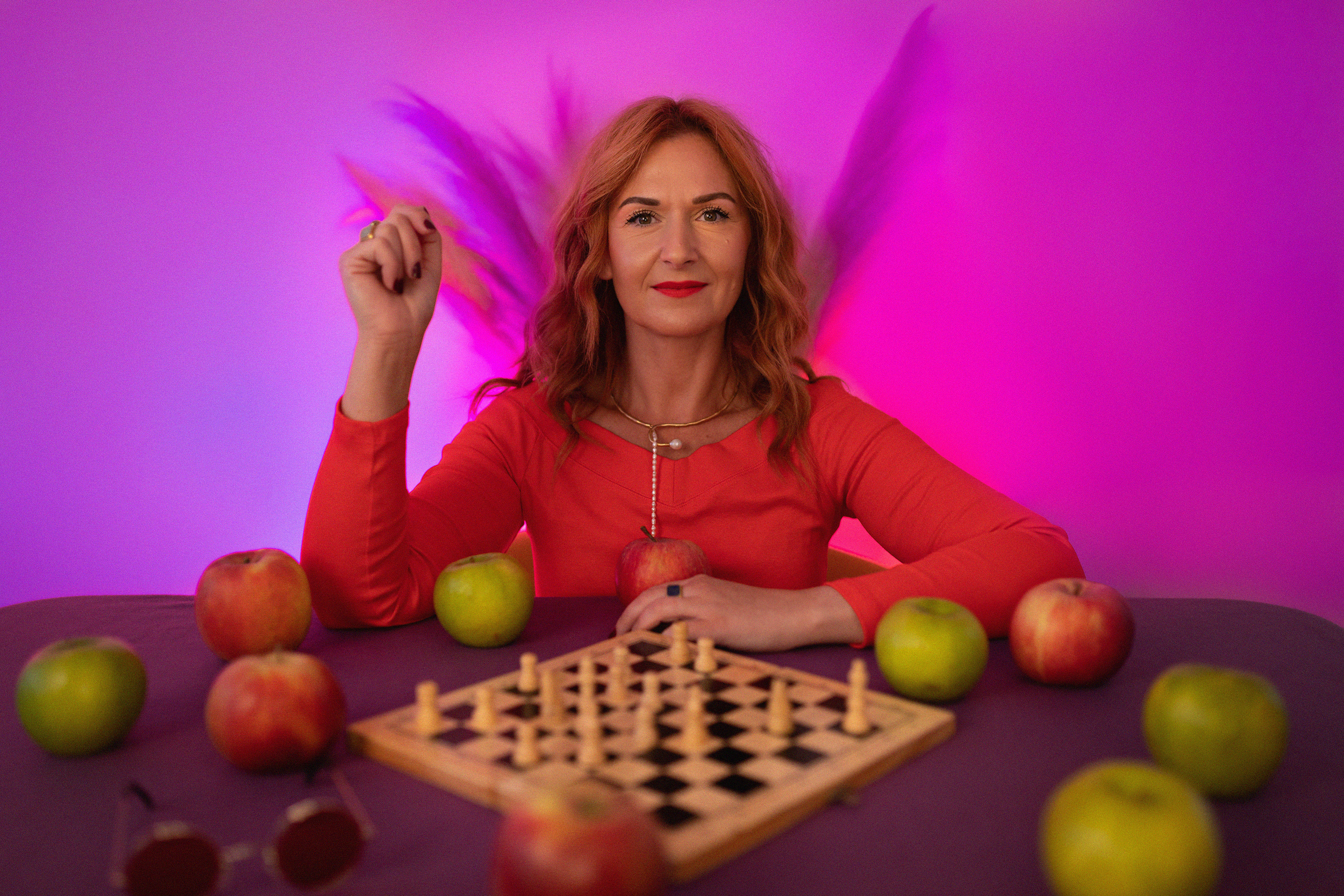 A woman with wavy red hair, wearing a red top and layered necklaces, sits at a table with apples and a chessboard. She raises her right arm with a clenched fist and has a confident expression. The background is lit with purple and pink hues, and there are colorful feathers behind her.