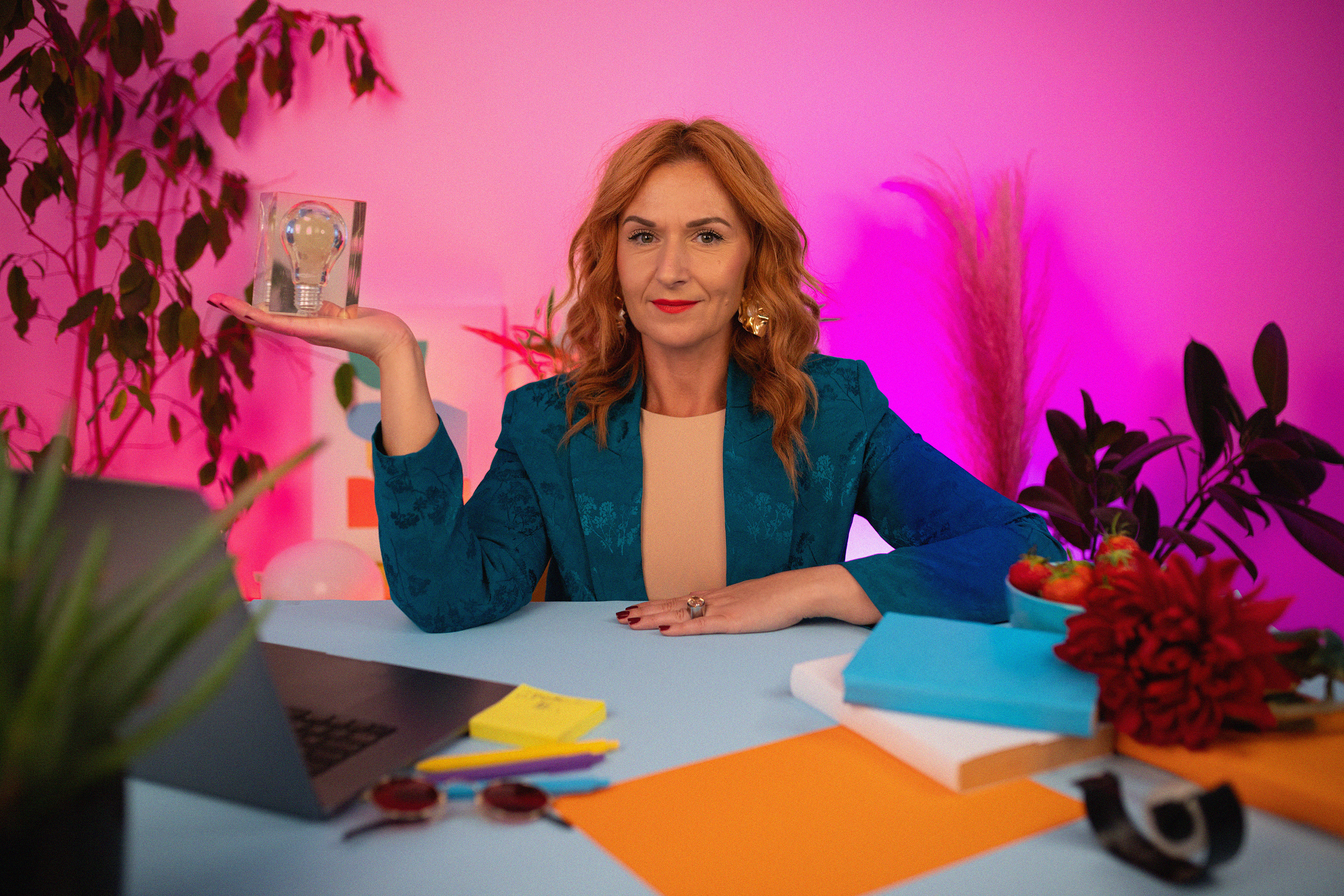 A woman with red hair, wearing a blue blazer, sitting at a colorful desk surrounded by plants and colorful stationery, holding a glass block with a light bulb inside, against a pink-lit background.