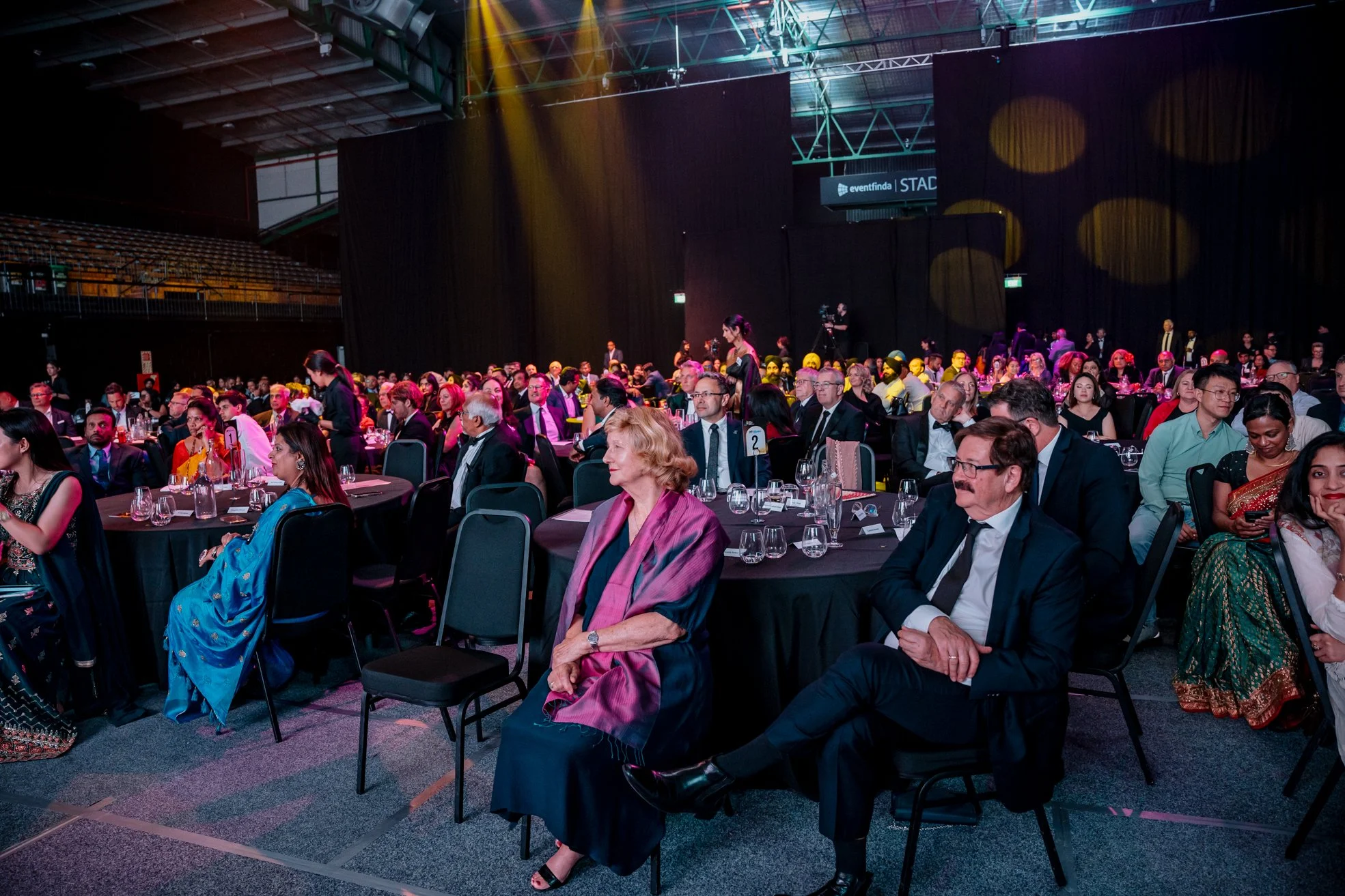 A large audience seated at round tables in a conference hall or banquet setting, with a stage in the background, some people in traditional Indian attire, and a black curtain backdrop.