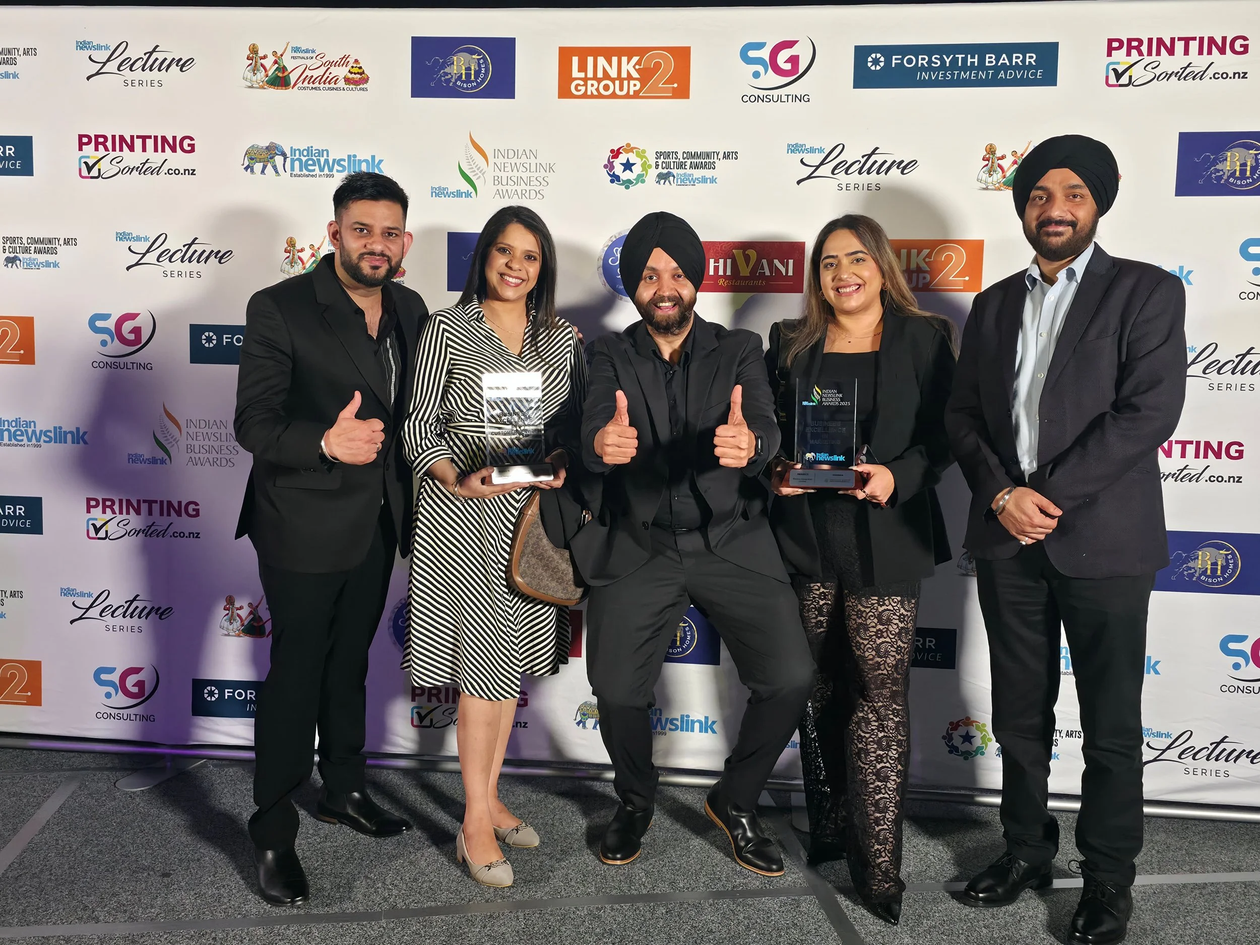 Group of five people standing on stage, holding awards, at an event with a backdrop featuring various logos and sponsors, smiling and posing for a photo.