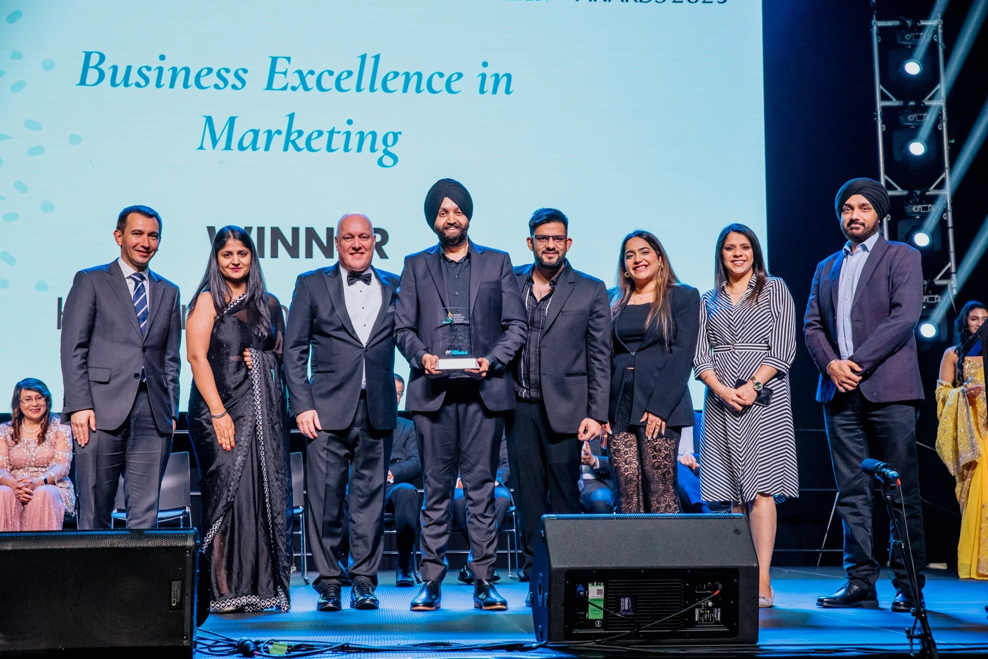 Group of people standing on stage during an award ceremony, with a large screen behind them displaying 'Business Excellence in Marketing' and 'WINNER' in bold letters.