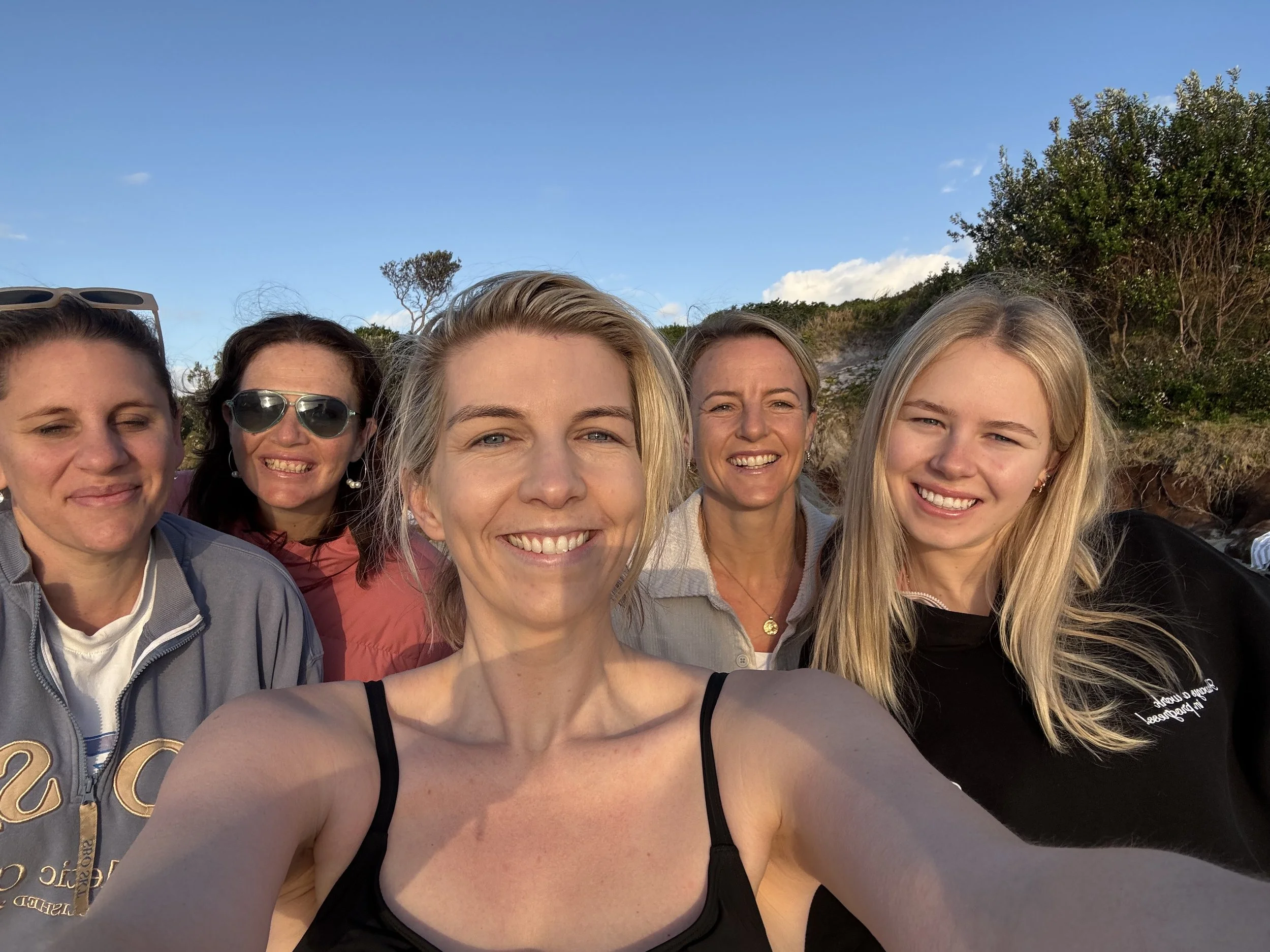 Five Wollongong women smiling and taking a selfie outdoors on a sunny day with green bushes and a blue sky in the background.