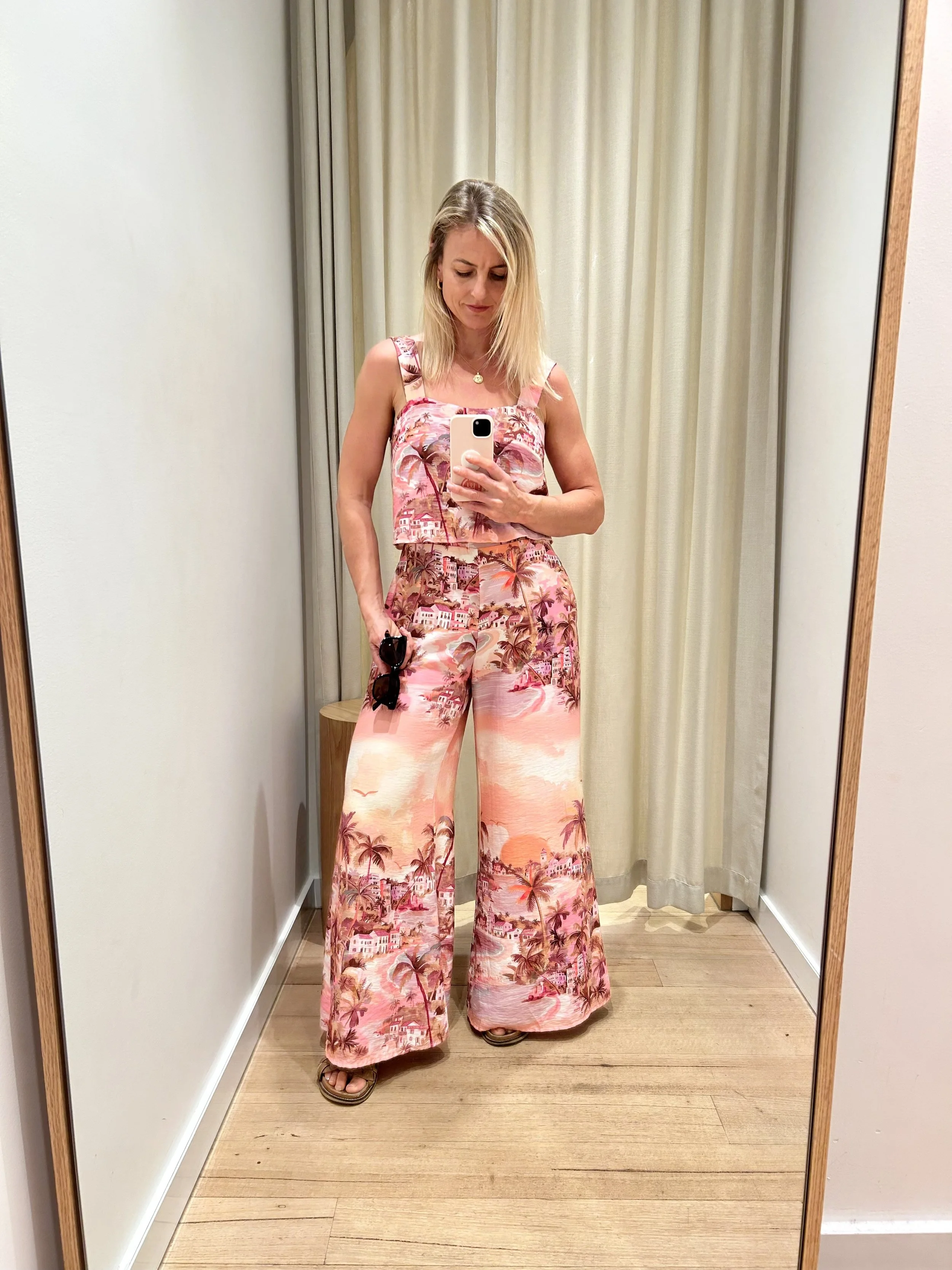 Woman in a pink tropical print top and wide-leg pants taking a mirror selfie in a fitting room.