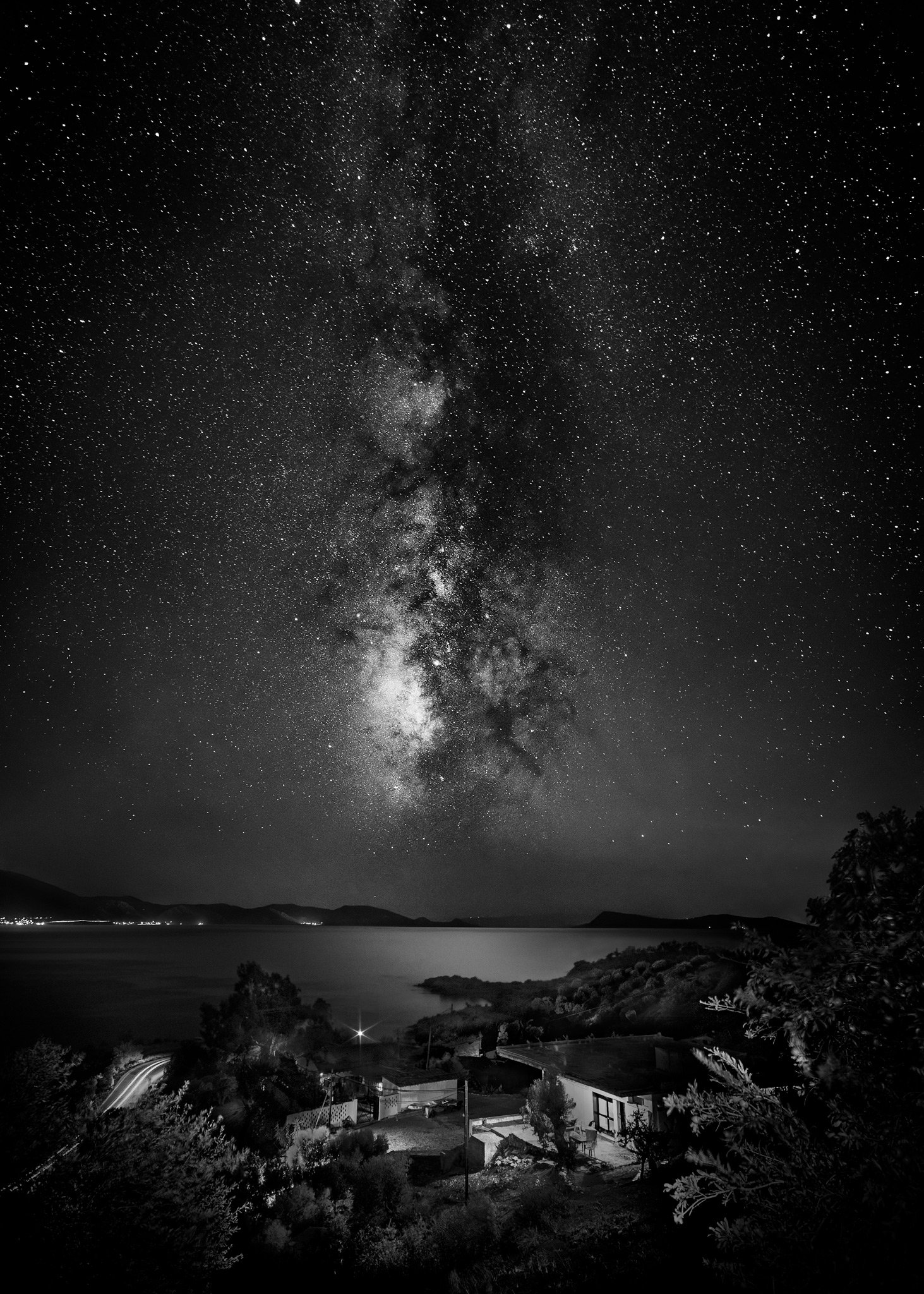 Black and white photo of starry night sky with the Milky Way galaxy, overlooking a lake with houses and trees in the foreground.