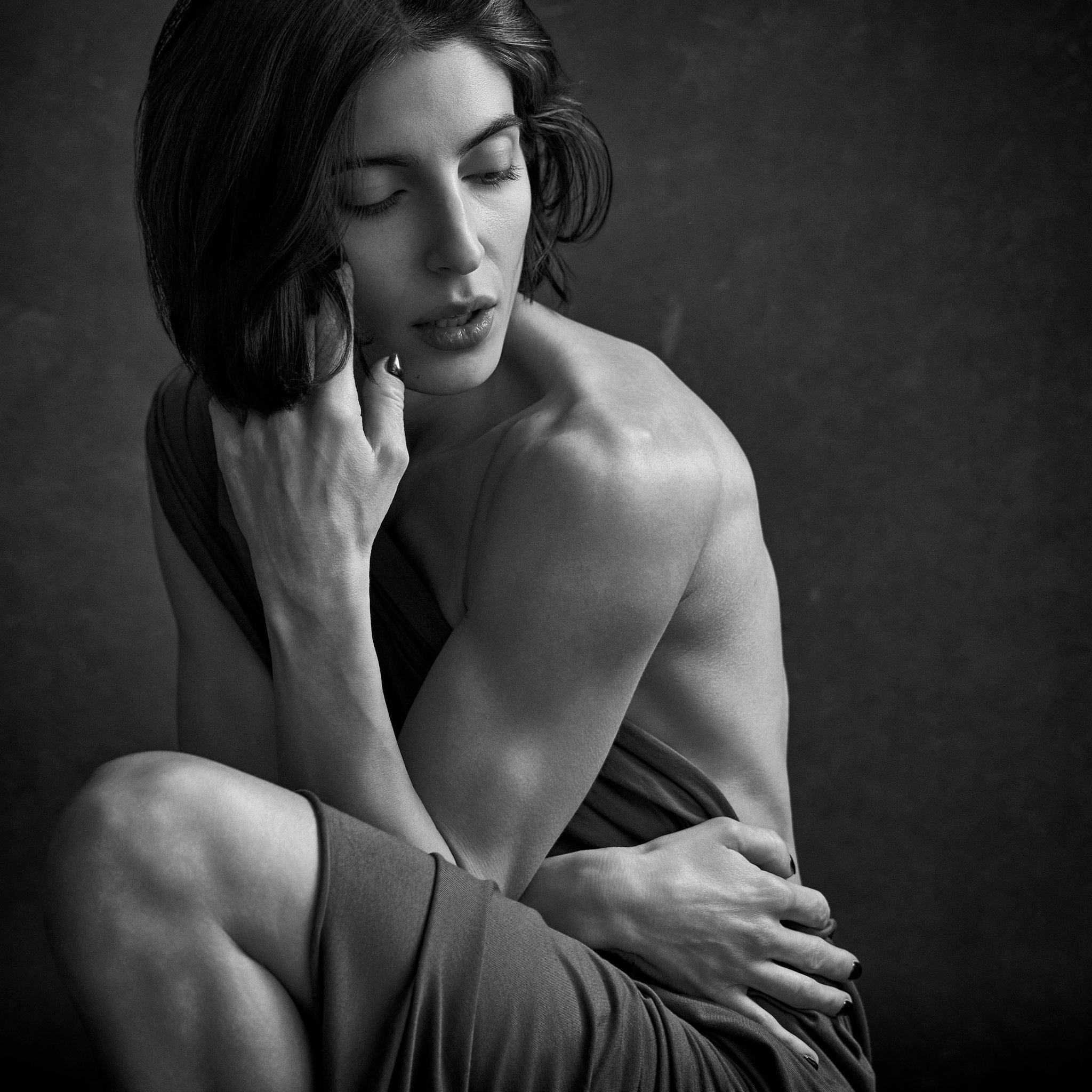 Black and white close-up photo of a woman with her eyes closed, resting her face against her hand and shoulder, with short wavy hair.