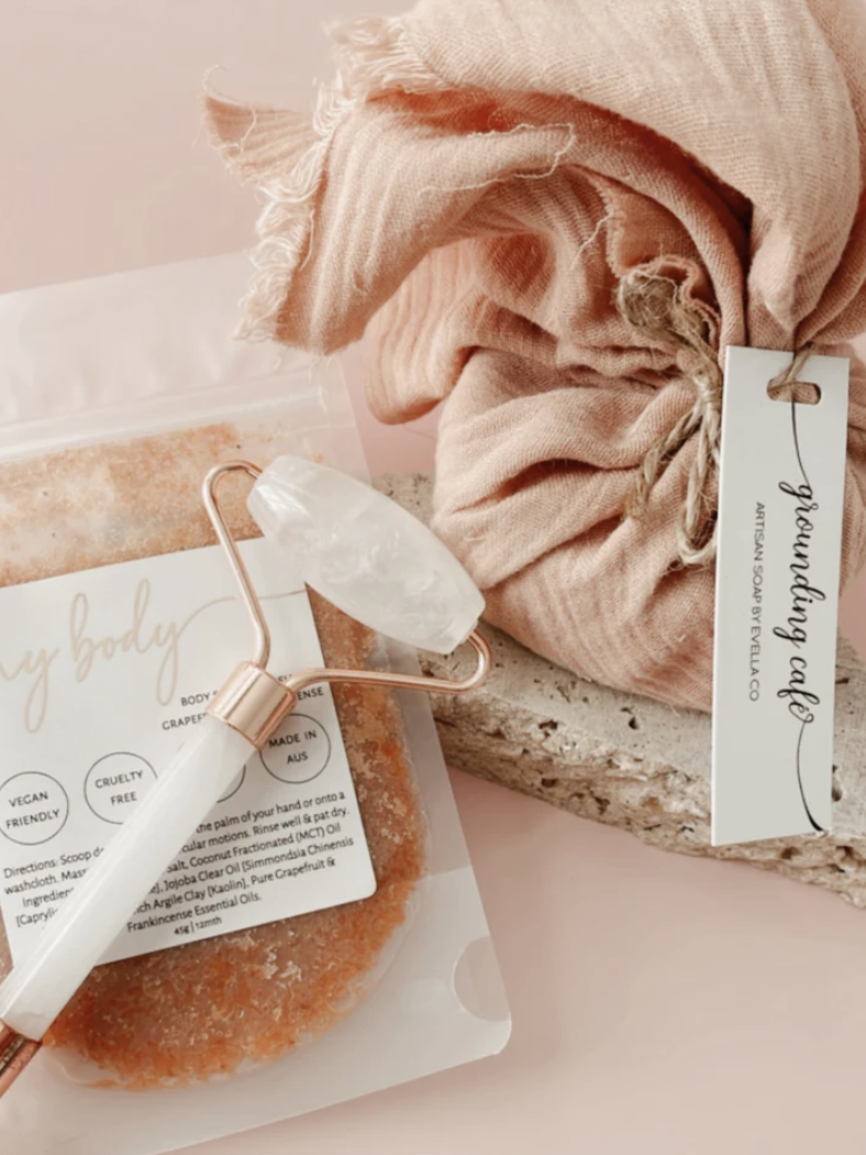 A package of artisan soap labeled 'My Body' with a pink and white rose quartz facial roller resting on it, and a pink cloth tied with a tag that reads 'Heading Café'.