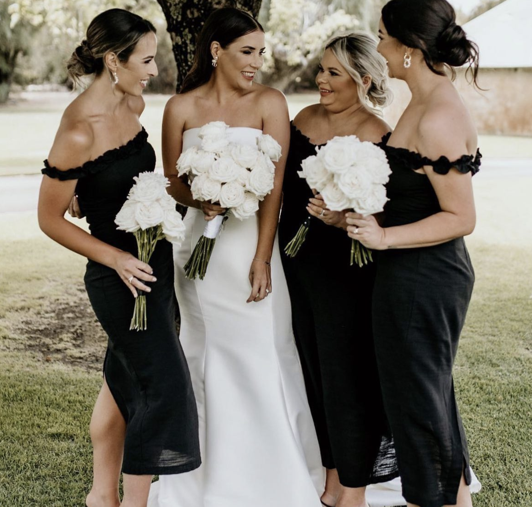 A bride in a white wedding dress holding a bouquet of white roses, standing with three women in black dresses holding matching bouquets, outdoors on grass with trees in the background.