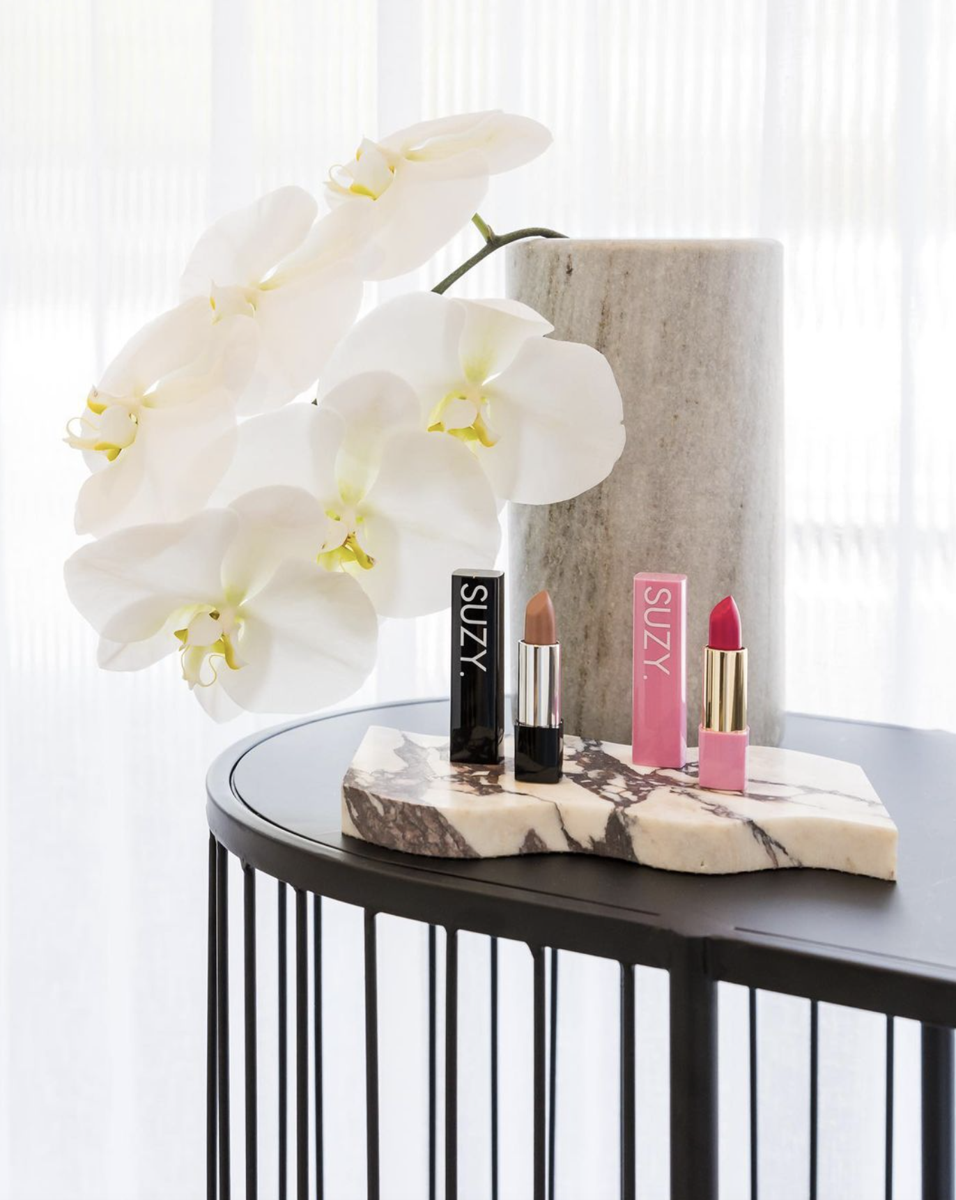 Beauty display with two lipsticks, a potted orchid plant, and a marble slab on a black metal table near a sheer white curtain.