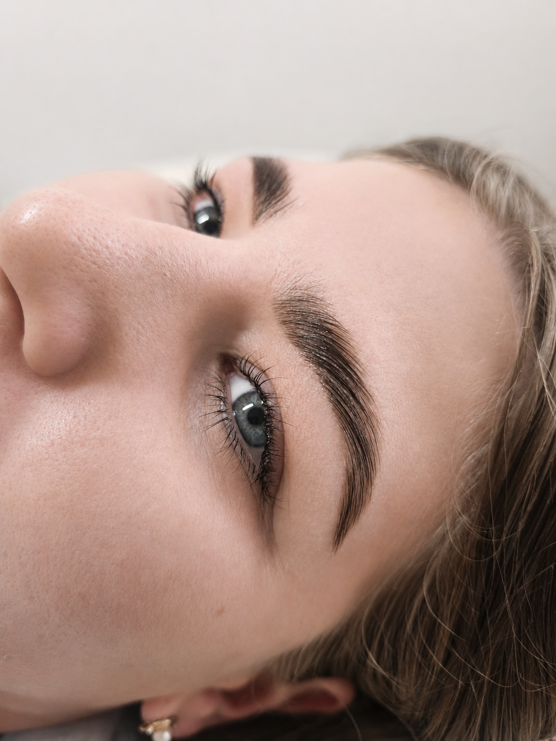 Close-up photo of a young woman with fair skin and freckles, lying down on a beige chair, with dark brown hair, thick arched eyebrows, light makeup, and a small gold earring.