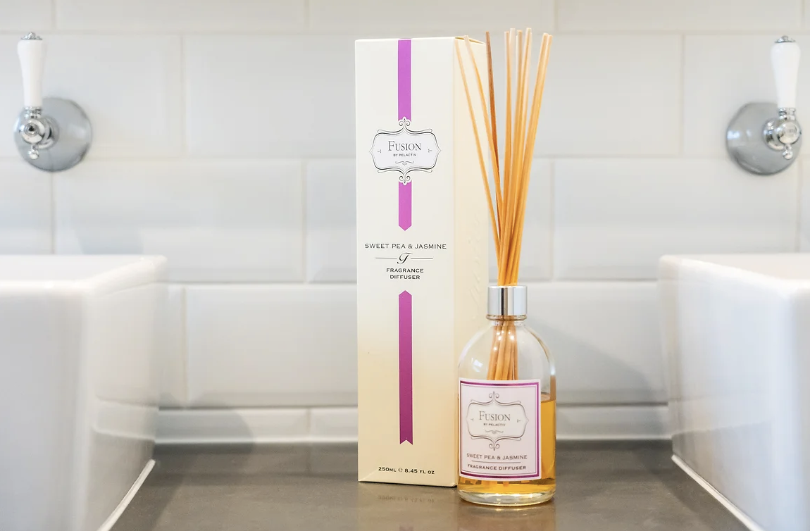 A fragrance diffuser with reed sticks and packaging labeled "Fusion by Pelactiv" and "Sweet Pea & Jasmine" on a bathroom countertop with white tile walls.