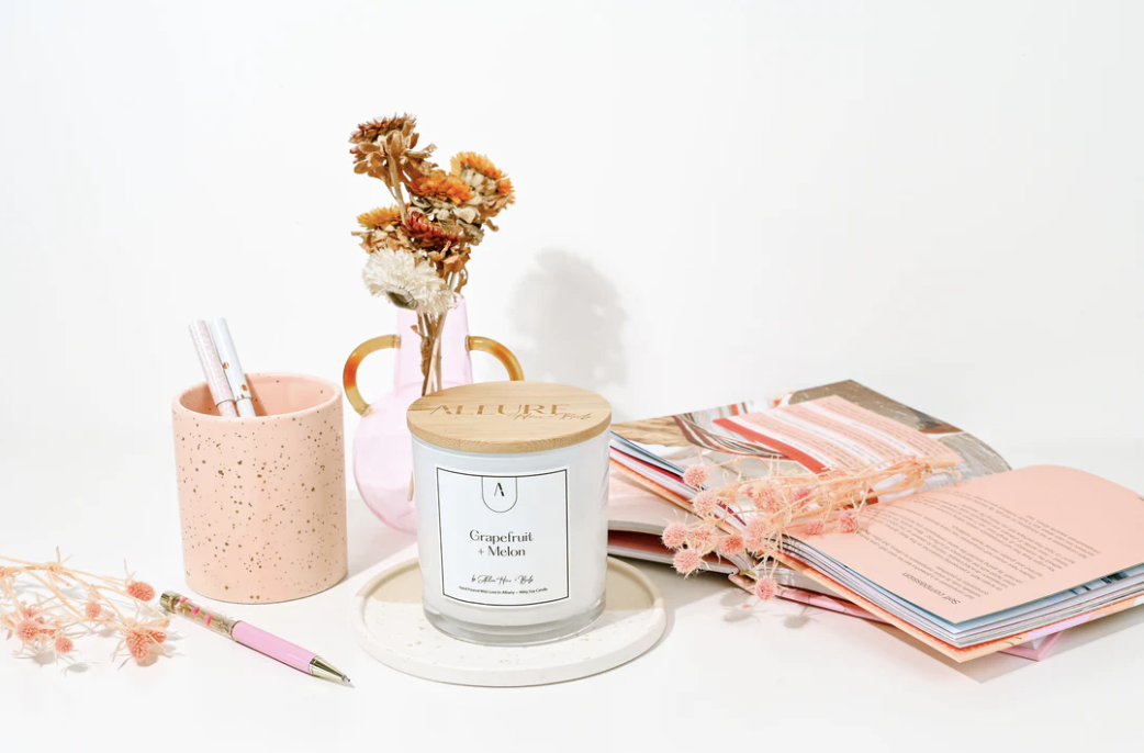 Pink office desk items including a speckled pink cup with pens, a pink vase with dried flowers, a white candle with a wooden lid labeled 'Grapefruit + Melon,' an open magazine with pink accents, and pink dried flowers arranged across the desk.