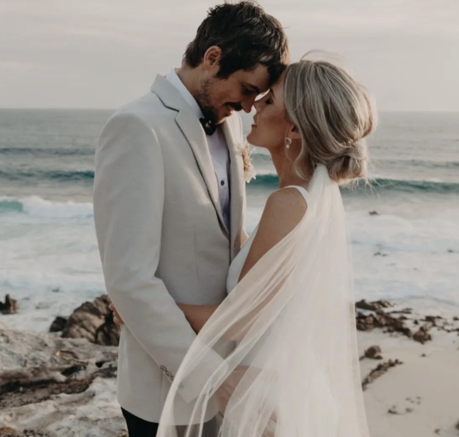 A bride and groom standing close together on the beach, with the ocean in the background as the sun sets, about to kiss.