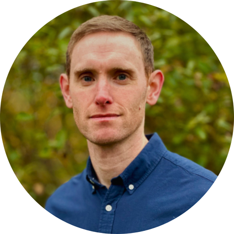 Portrait of a man outdoors with a blurred green foliage background, wearing a blue button-down shirt.