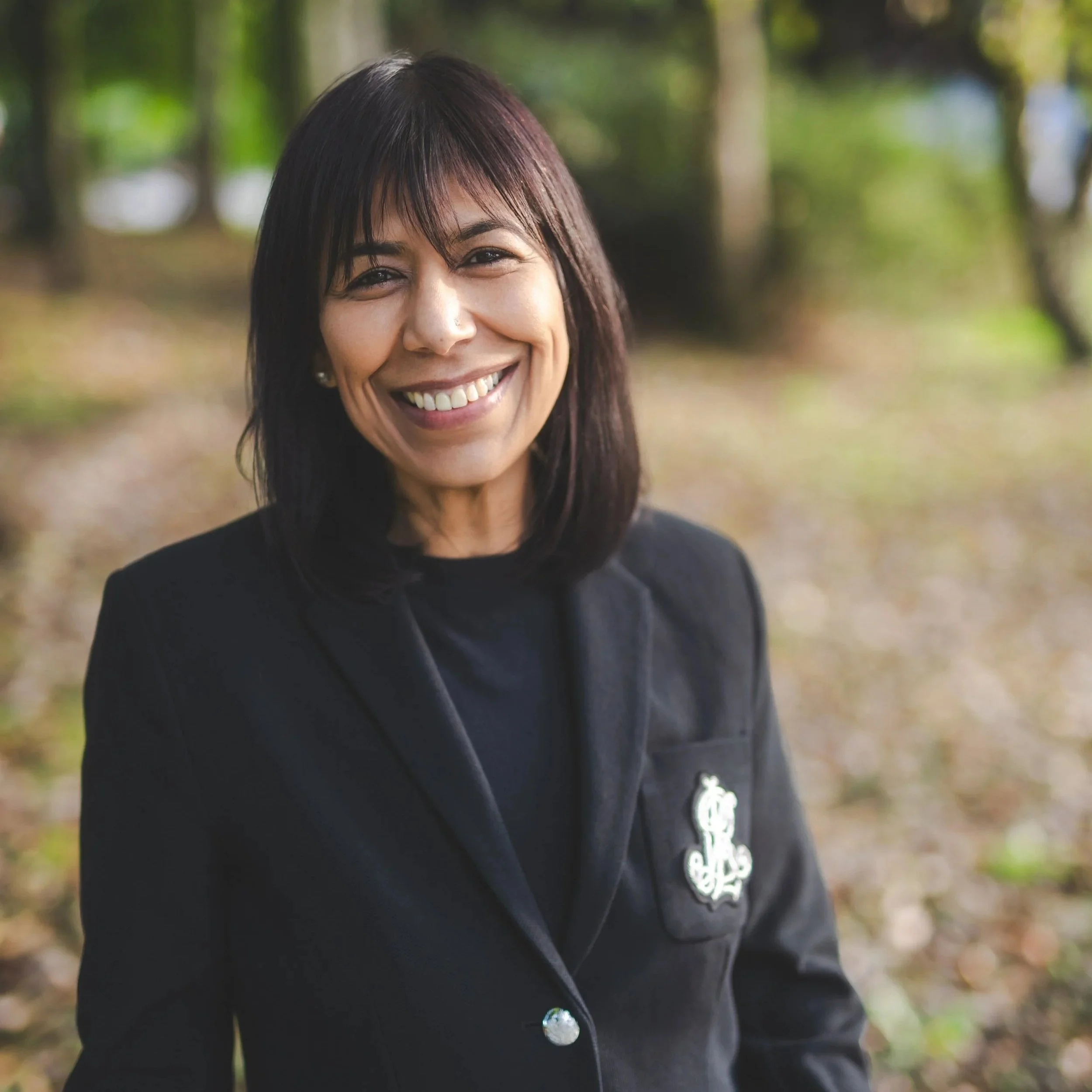 A woman with black hair, wearing a black top with white accents, smiling with arms crossed, standing outdoors with green trees in the background.