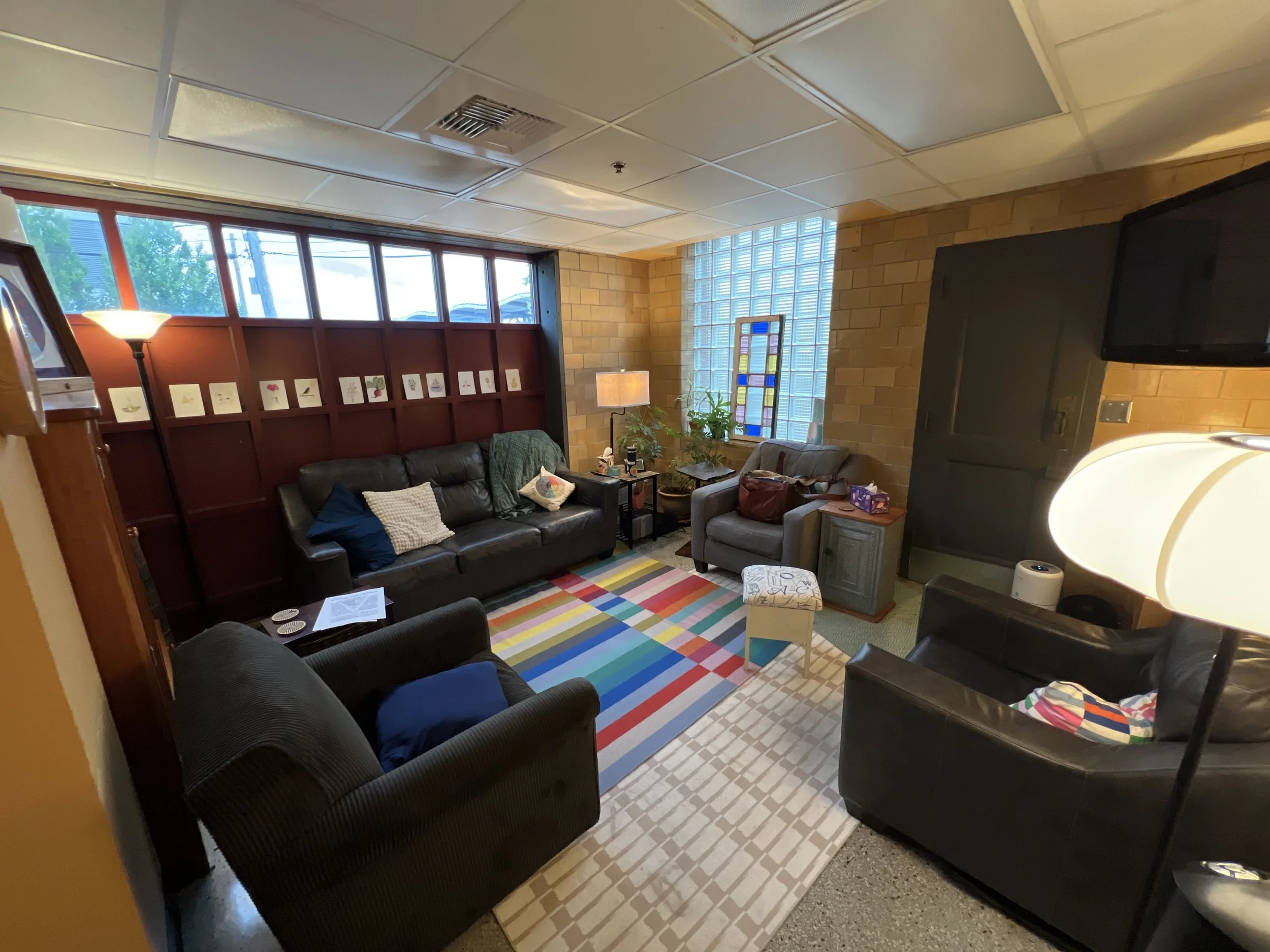 Cozy living room with three black leather sofas, a patterned rug, a side table with a lamp, plants by the window, and colorful artwork on the wall.