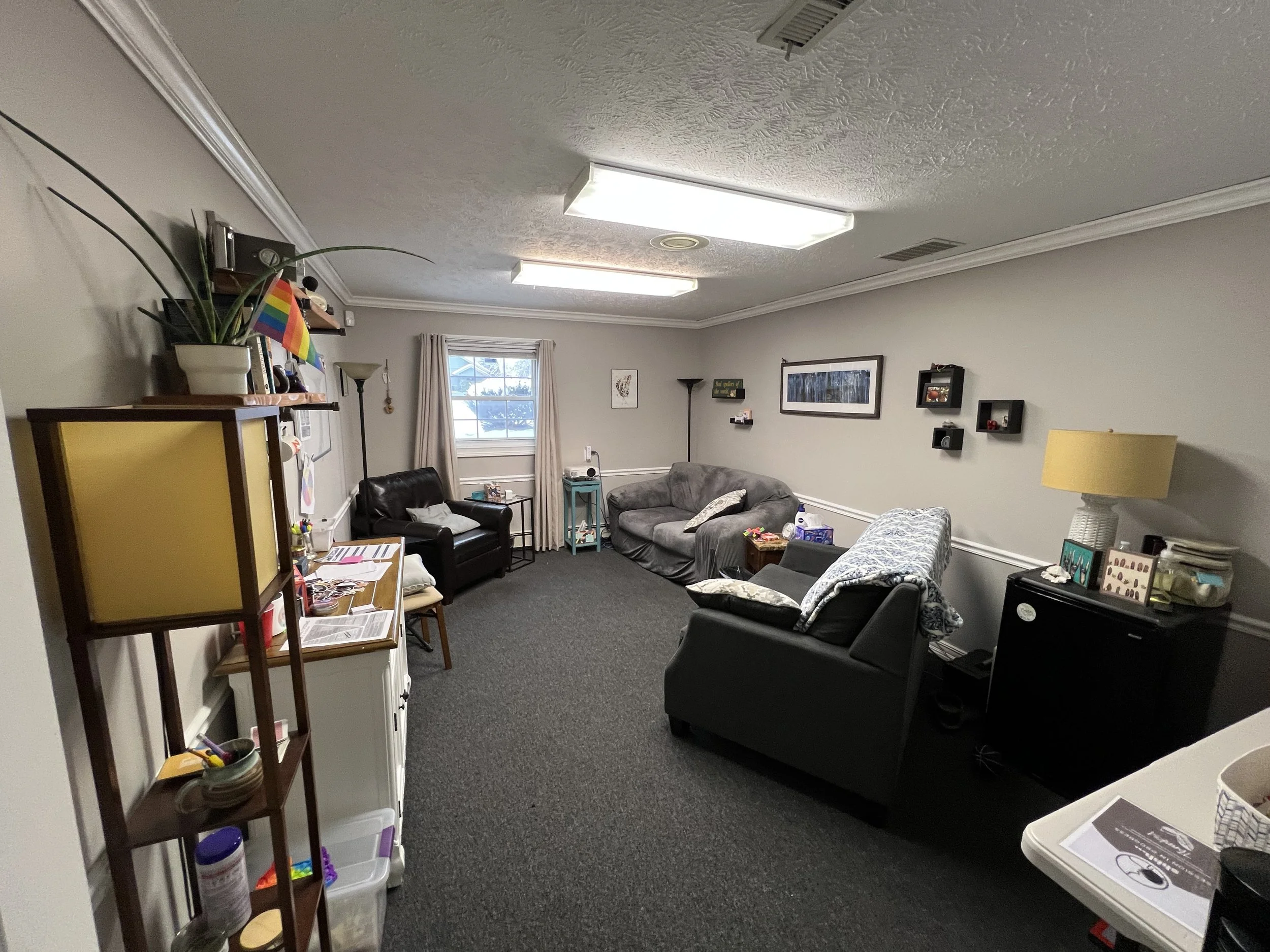 A living room with three sofas, a window with beige curtains, wall decorations, a lamp, a standing lamp, and a small table.