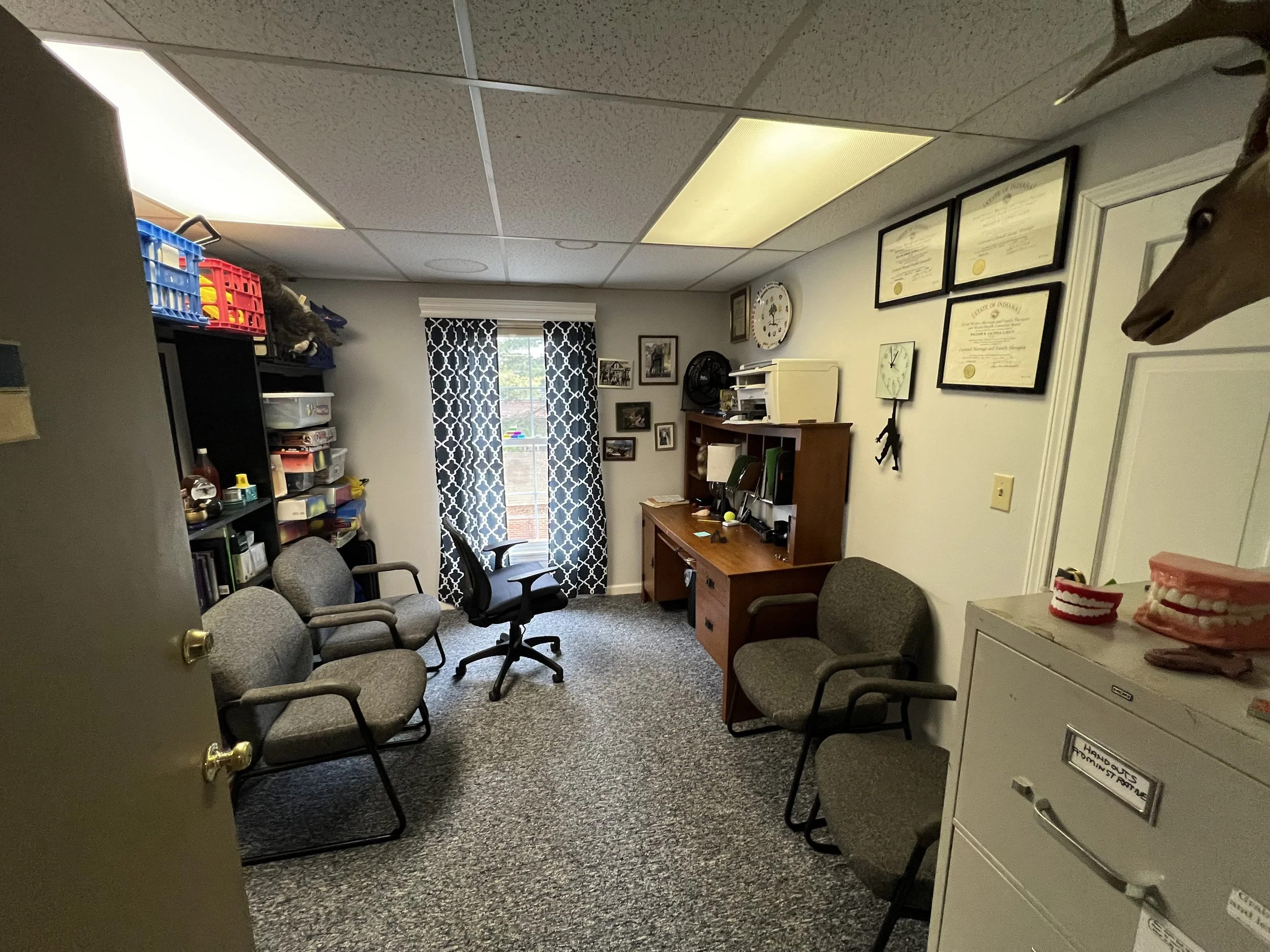 An office room with beige walls and ceiling, containing a wooden desk with a computer, chairs, bookshelves filled with books and supplies, framed certificates on the wall, and a window with patterned curtains.