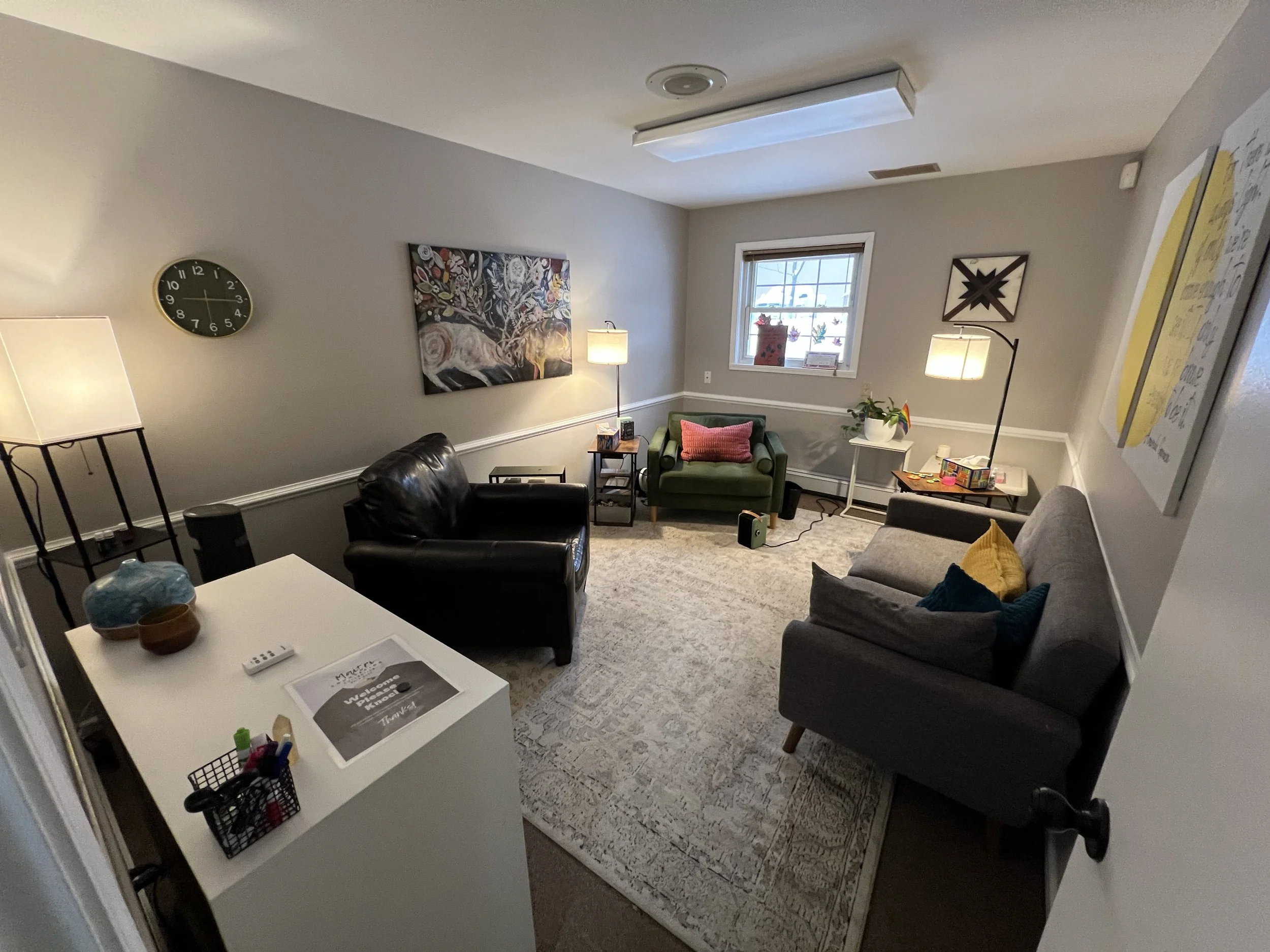 A cozy living room with a gray sofa, green armchair, black leather armchair, and a small dining table. The room is decorated with colorful pillows, artwork on the walls, and several lamps providing warm lighting. There's a window with a view outside and various decorative items on tables and shelves.