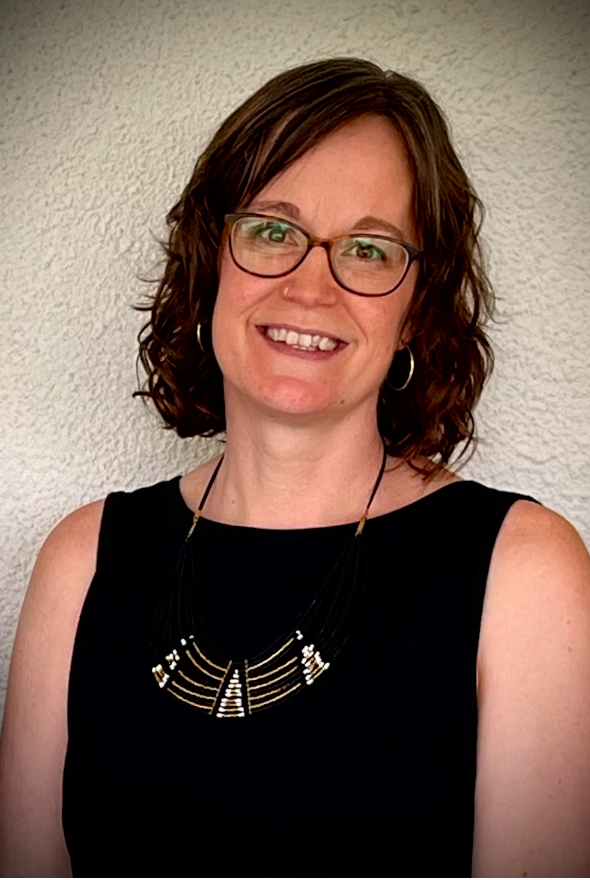 A smiling woman with shoulder-length curly brown hair, wearing glasses, hoop earrings, a black sleeveless top, and a layered black and gold necklace, standing in front of a white textured wall.